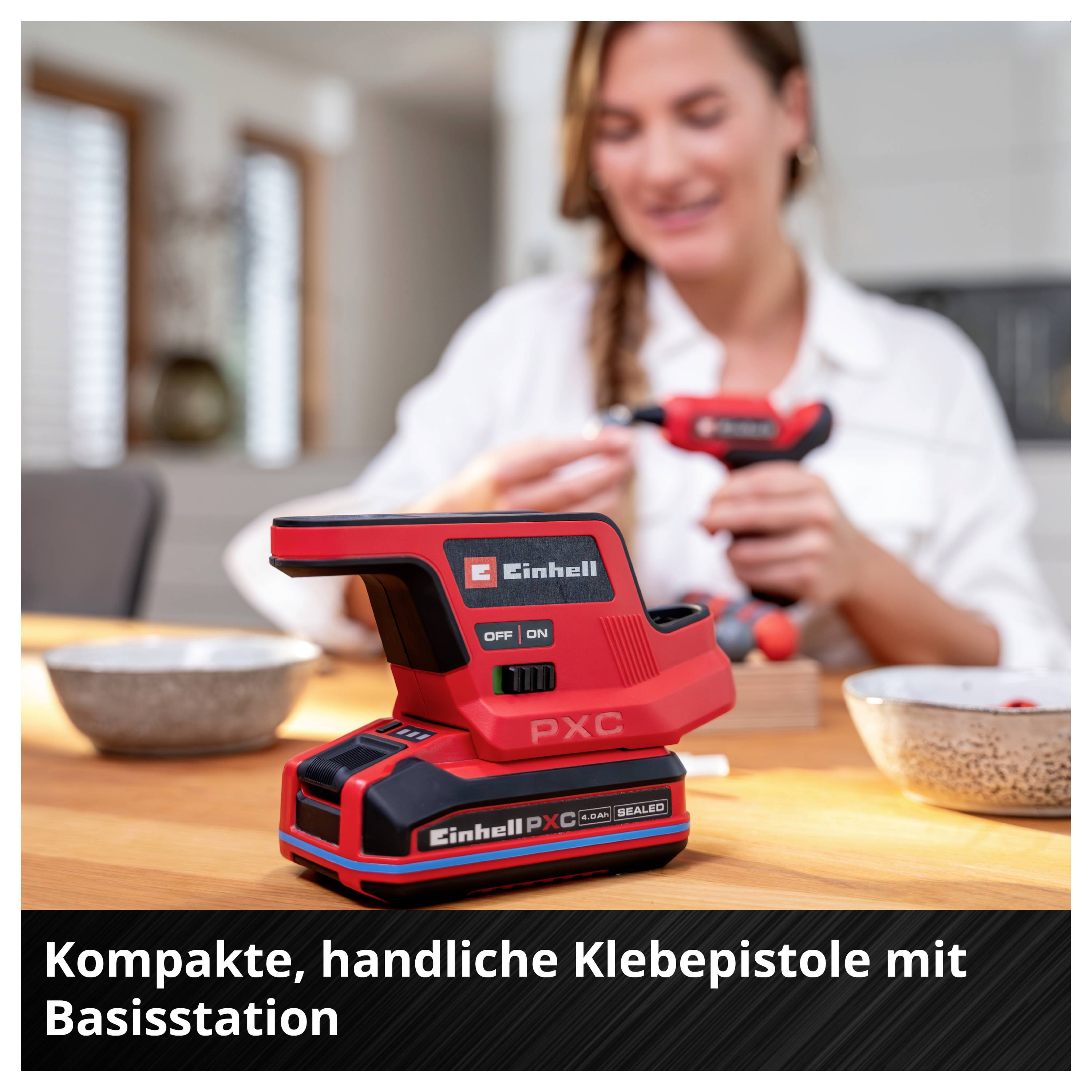 Kompakte rote Einhell-Pistolen-Klebepistole mit Basisstation auf einem Tisch, im Hintergrund eine Frau beim Arbeiten. Text: 'Kompakte, handliche Klebepistole mit Basisstation'