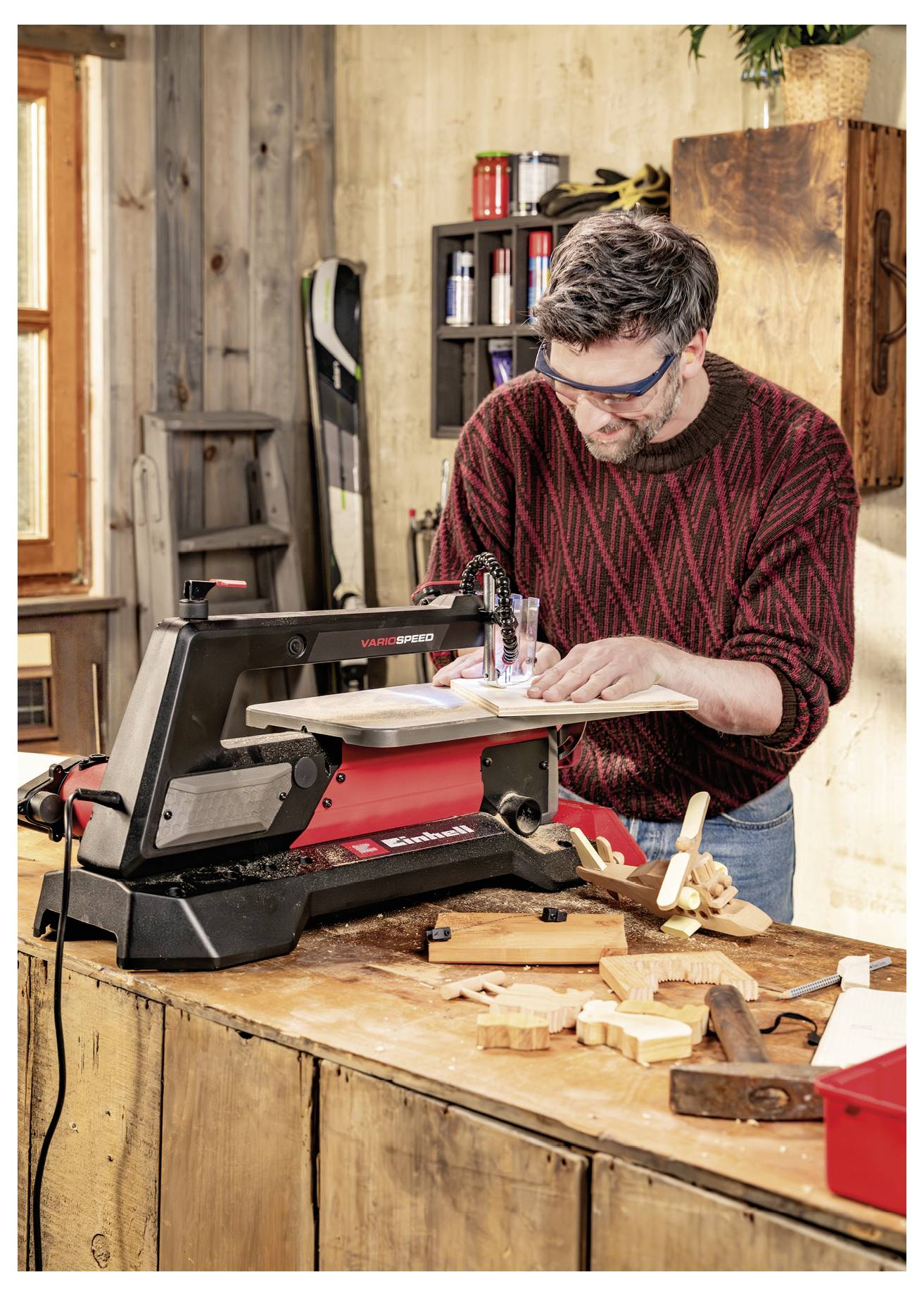 Mann benutzt eine Dekupierssäge auf einer Holzwerkbank in einer Werkstatt und schneidet Holzstücke, während er eine Schutzbrille trägt.
