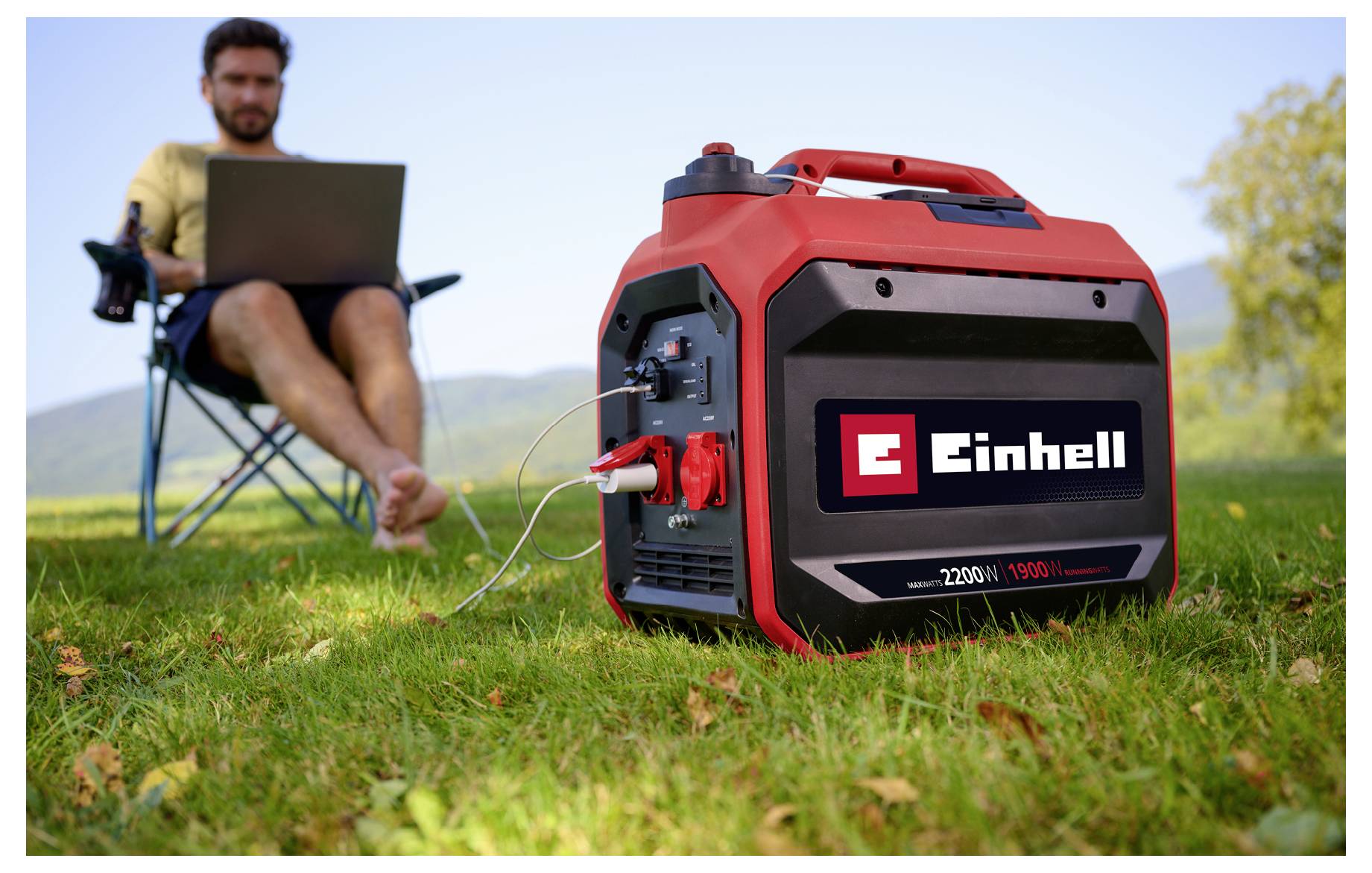 Tragbarer Einhell-Generator auf Gras, der einen Laptop mit Strom versorgt, mit einem Mann, der im Hintergrund sitzend den Laptop im Freien nutzt.