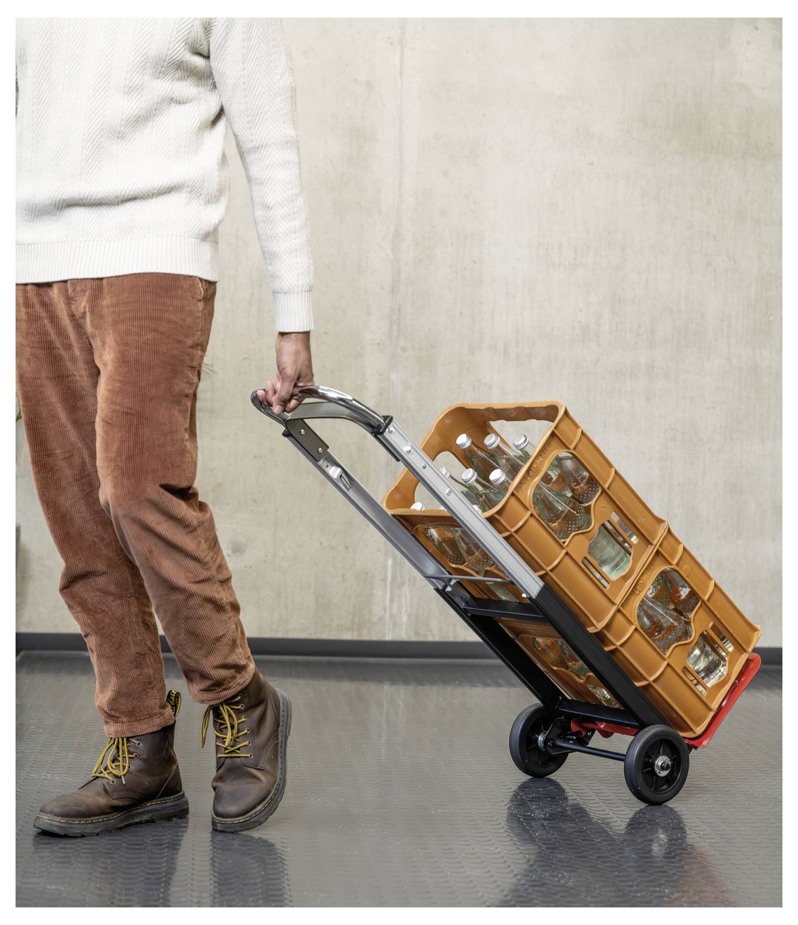 Person in weißem Pullover und brauner Hose zieht einen Handwagen, der mit orangefarbenen Getränkekisten und Glasflaschen beladen ist.