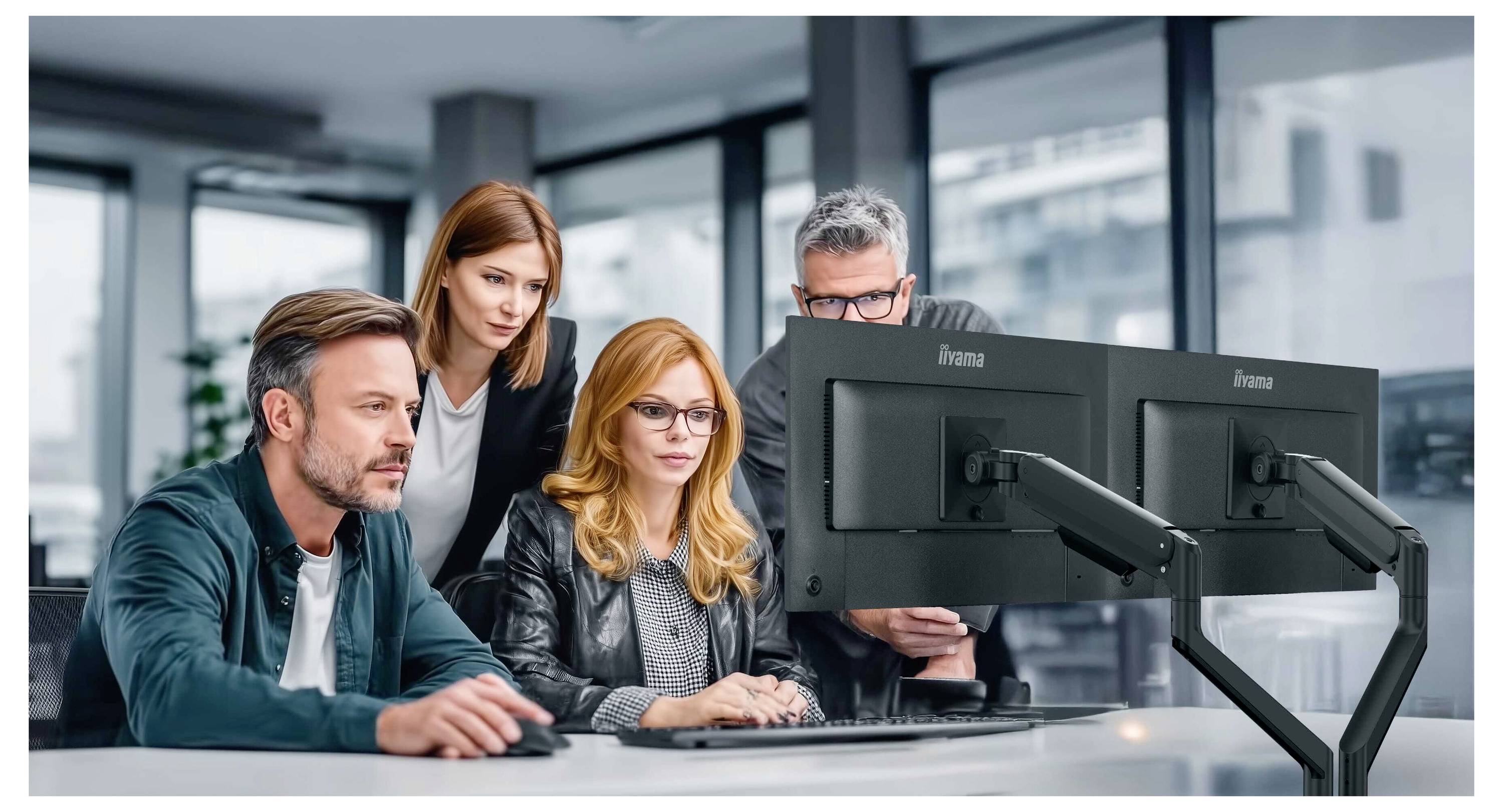 Vier Kollegen studieren Daten auf dualen Computermonitoren in einem Büro, wobei zwei sitzen und zwei hinter ihnen stehen.