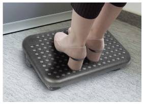 Person standing on a black textured Fußstütze mit erhobenen Fersen, wahrscheinlich zur Unterstützung oder zum Komfort am Schreibtisch.