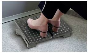 Füße in rosa hochhackigen Sandalen drücken auf eine Fußmassage-Rolle auf einem grau teppichbelegten Boden.