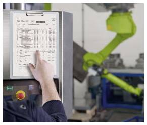Ein Arbeiter bedient ein Touchscreen-Bedienfeld, während ein grüner Industrieroboterarm im Hintergrund in einer Fabrik arbeitet.