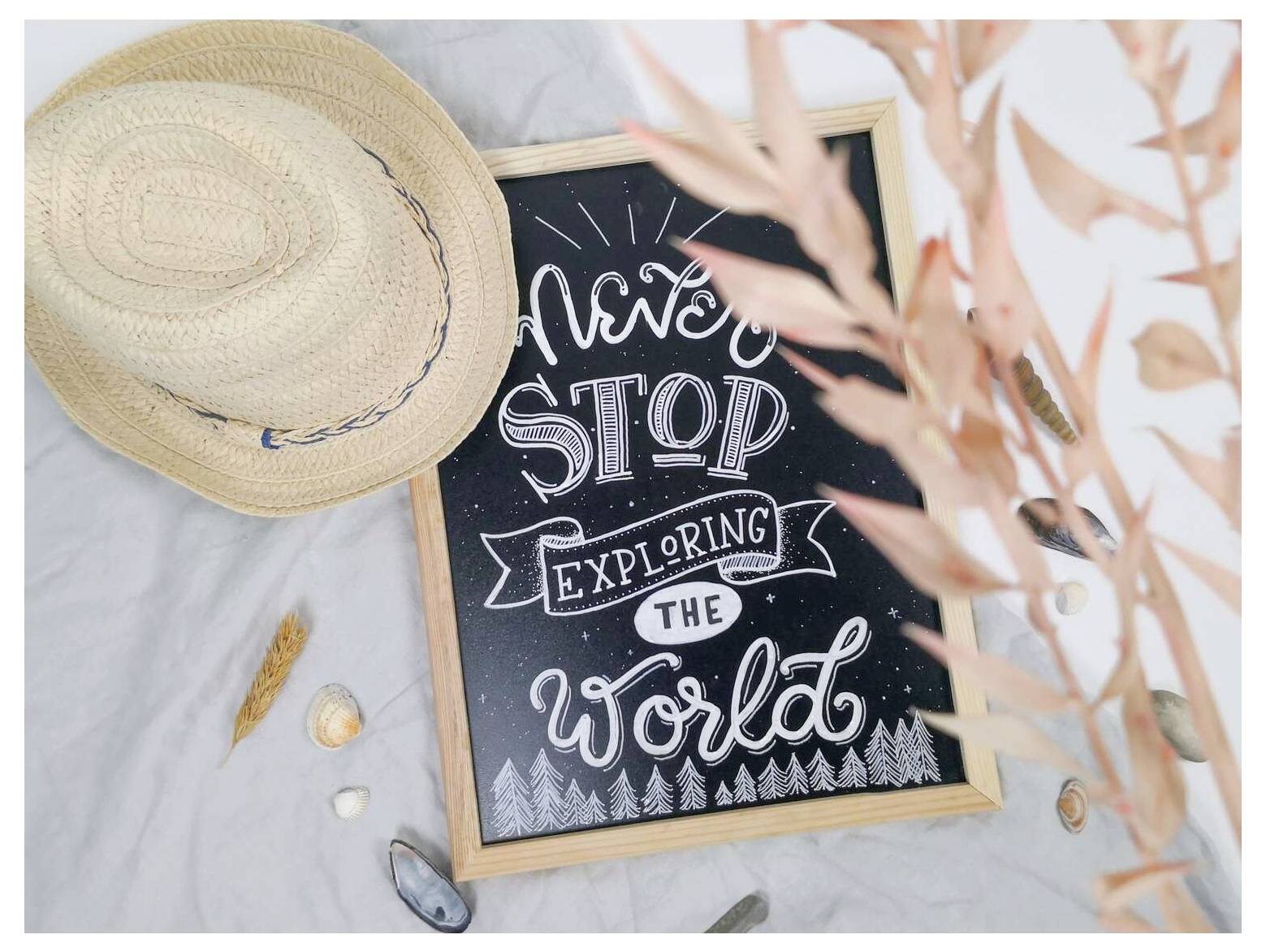 Schreibtafel in einer Strandszene zeigt die Aufschrift „Never STOP EXPLORING THE World" neben einem Strohhut und Muscheln.