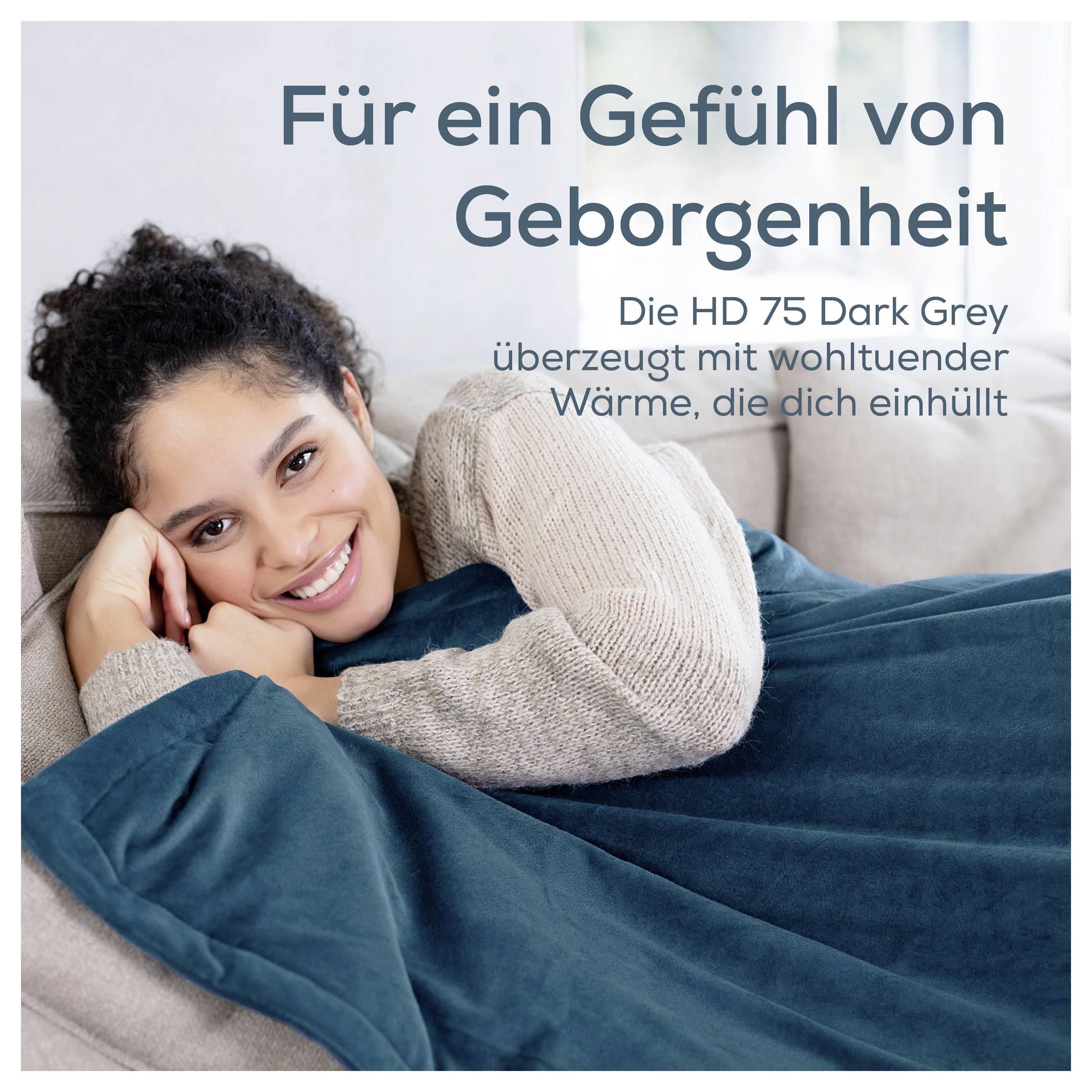 Lächelnde Frau liegt eingekuschelt unter einer dunkelgrauen Decke; Text: 'Für ein Gefühl von Geborgenheit' und 'Die HD 75 Dark Grey ... Wärme, die dich einhüllt'.