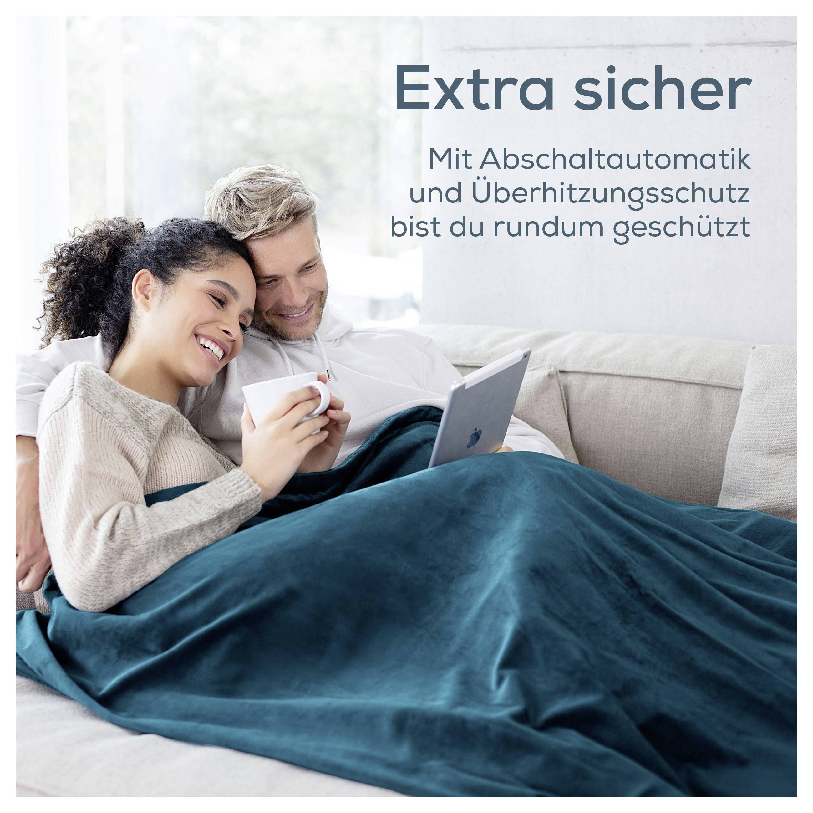 Paar kuschelt mit Laptop unter Decke auf Sofa; Text: 'Extra sicher' und 'Mit Abschaltautomatik und Überhitzungsschutz bist du rundum geschützt'.