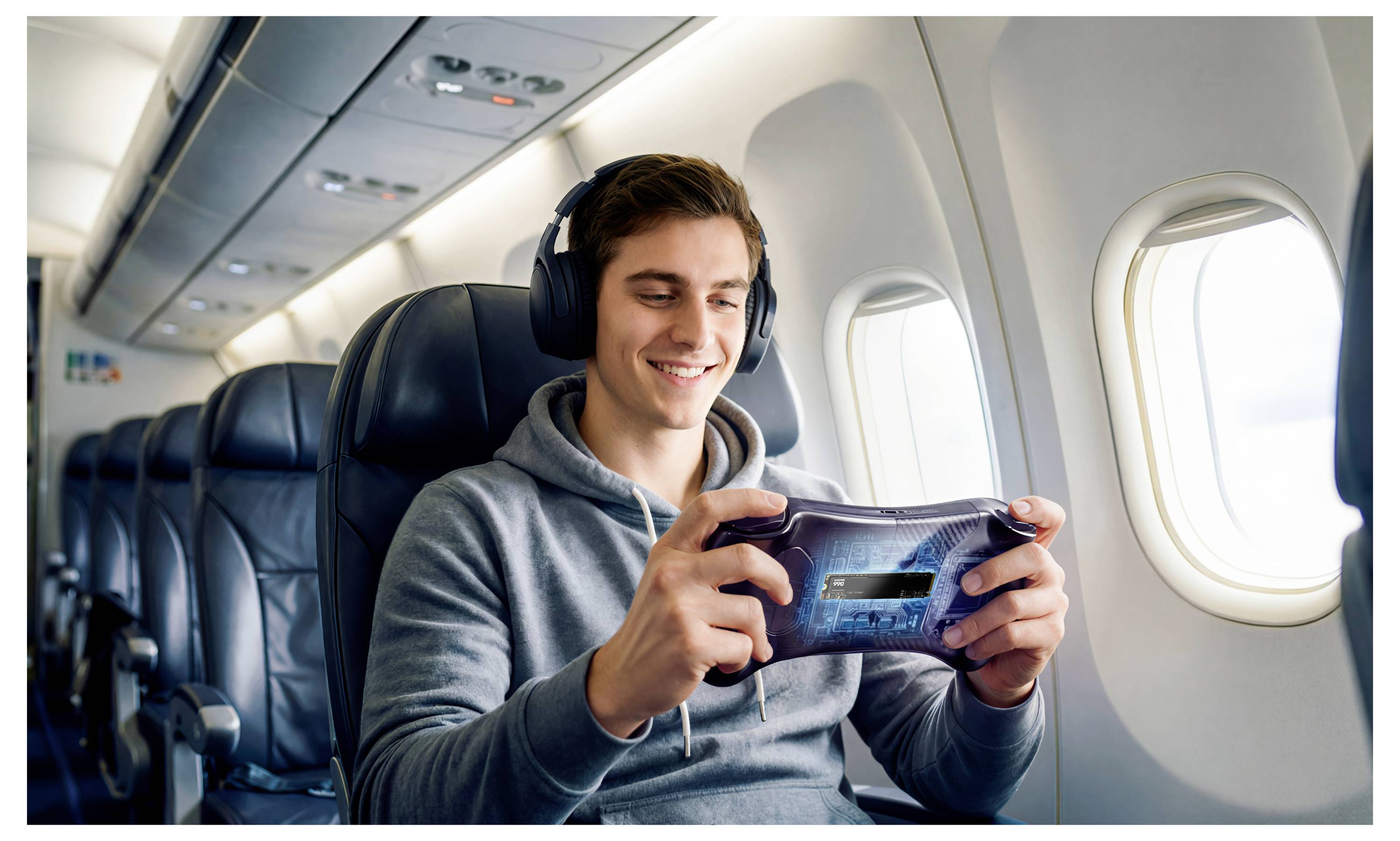 Ein junger Mann mit Kopfhörern lächelt, während er auf einem Flugzeugsitz am Fenster ein tragbares Spiel spielt.