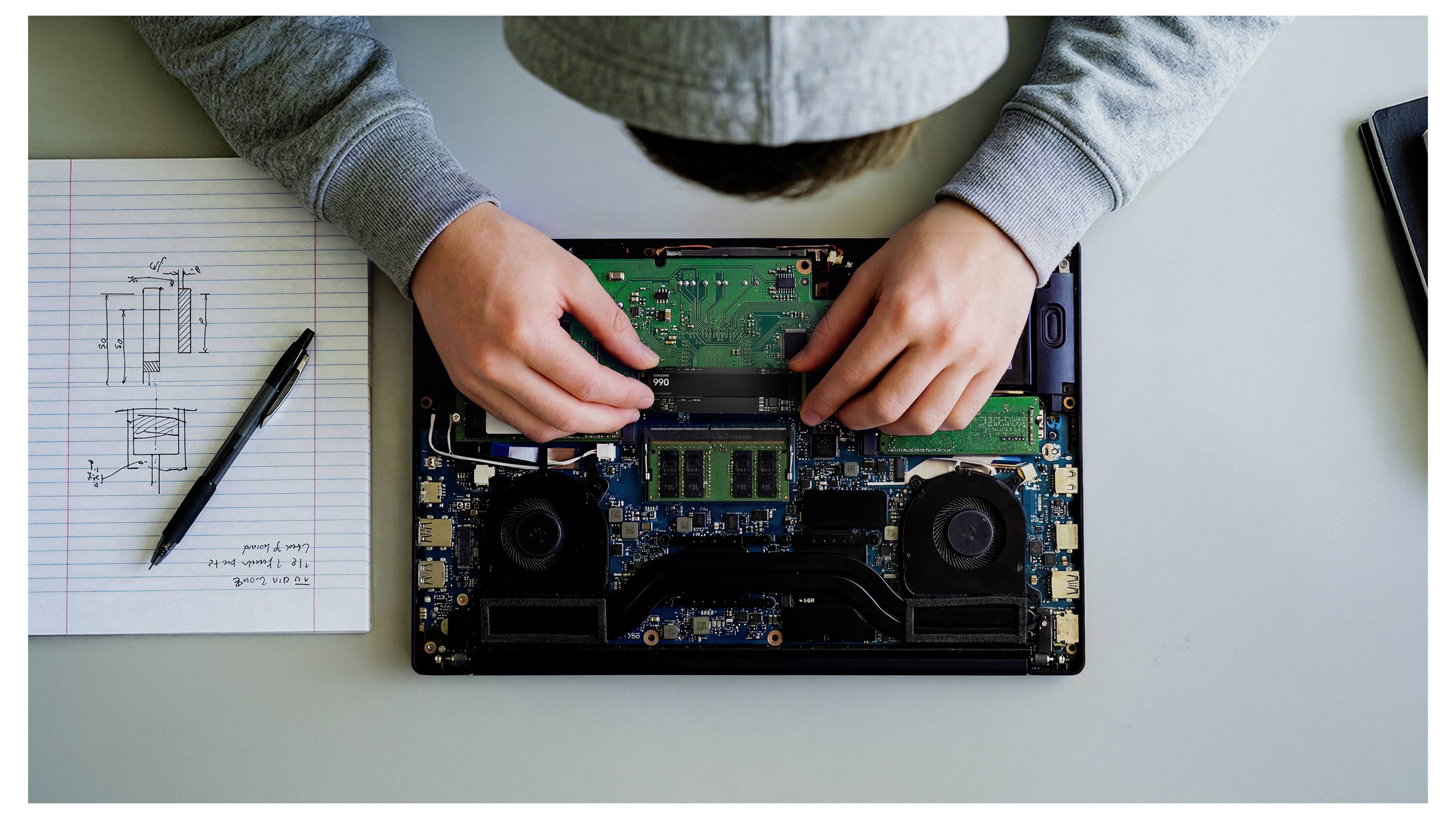 Person repariert einen Laptop mit entferntem Rückdeckel und hält die interne Platine auf einem Schreibtisch neben einem Notizbuch mit handgezeichneten technischen Notizen.