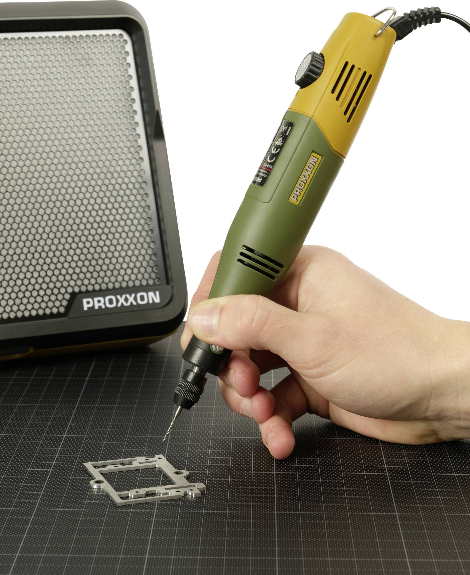 Hand hält ein PROXXON Rotationswerkzeug, um ein kleines Metallteil auf einer Schneidematte zu bohren, mit einer PROXXON Zubehörbox daneben.
