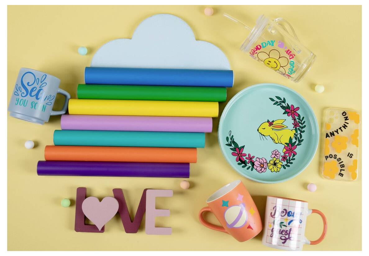 Bunter Flat-Lay-Arrangement mit Frühlings-Geschenkartikeln: Regenbogenstapel von Papierrollen, Becher, Tassen, ein Blumenteller, ein transparentes Trinkglas und ein LOVE-Schriftzug auf gelbem Hintergrund.