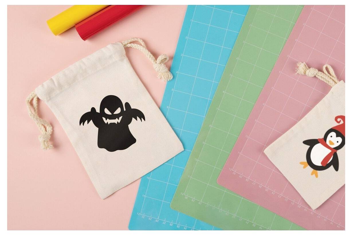 Zwei kleine Geschenktaschen mit Kordelzug auf farbigen Schneideteppichen, eine mit schwarzem Fledermaus- und Gespenster-Halloween-Design und eine mit einem Pinguin; rote und gelbe Marker liegen in der Nähe.