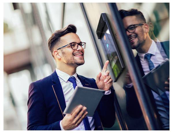 Geschäftsmann in einem Anzug bedient mit einem Tablet einen digitalen Touchscreen-Kiosk neben einem Glasfenster und lächelt dabei auf seine Spiegelung.