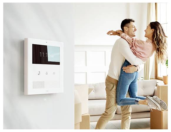 Ein Paar umarmt sich in einem hellen Wohnzimmer neben einem an der Wand montierten Heim-Sicherheits-/Gegensprechanlage-Panel, das die Zeit anzeigt.