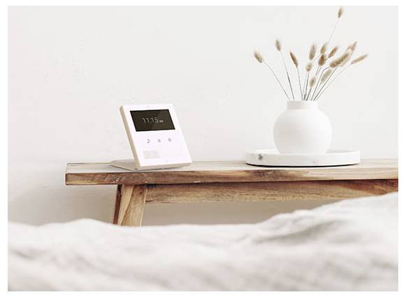 Minimalistisches Schlafzimmer mit einer Digitaluhr auf einem Holzregal und einer weißen Vase mit getrockneten Gräsern auf einem Tablett.