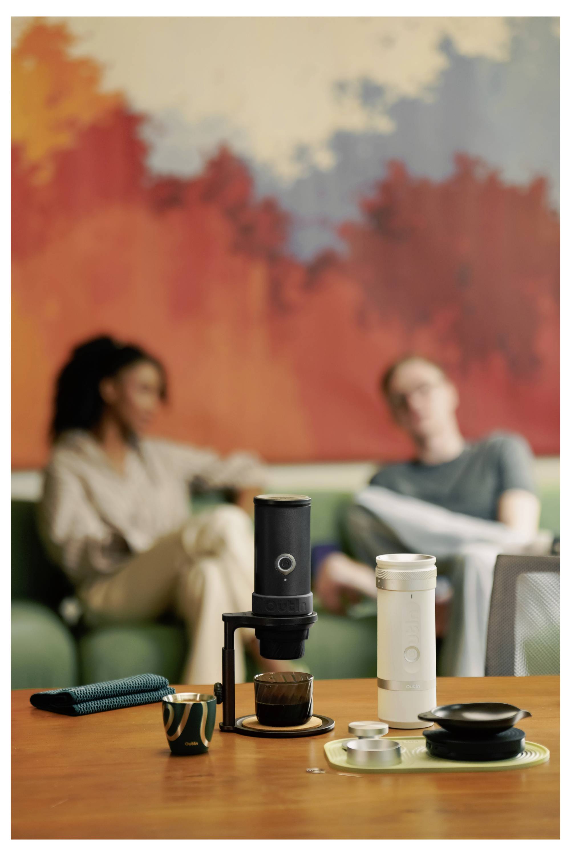 Kaffee-Brau-Setup auf einem Tisch mit einem schwarzen elektrischen Pour-Over-Dripper und einer weißen Reisebecher vor zwei unscharfen Personen, die auf einem Sofa sitzen