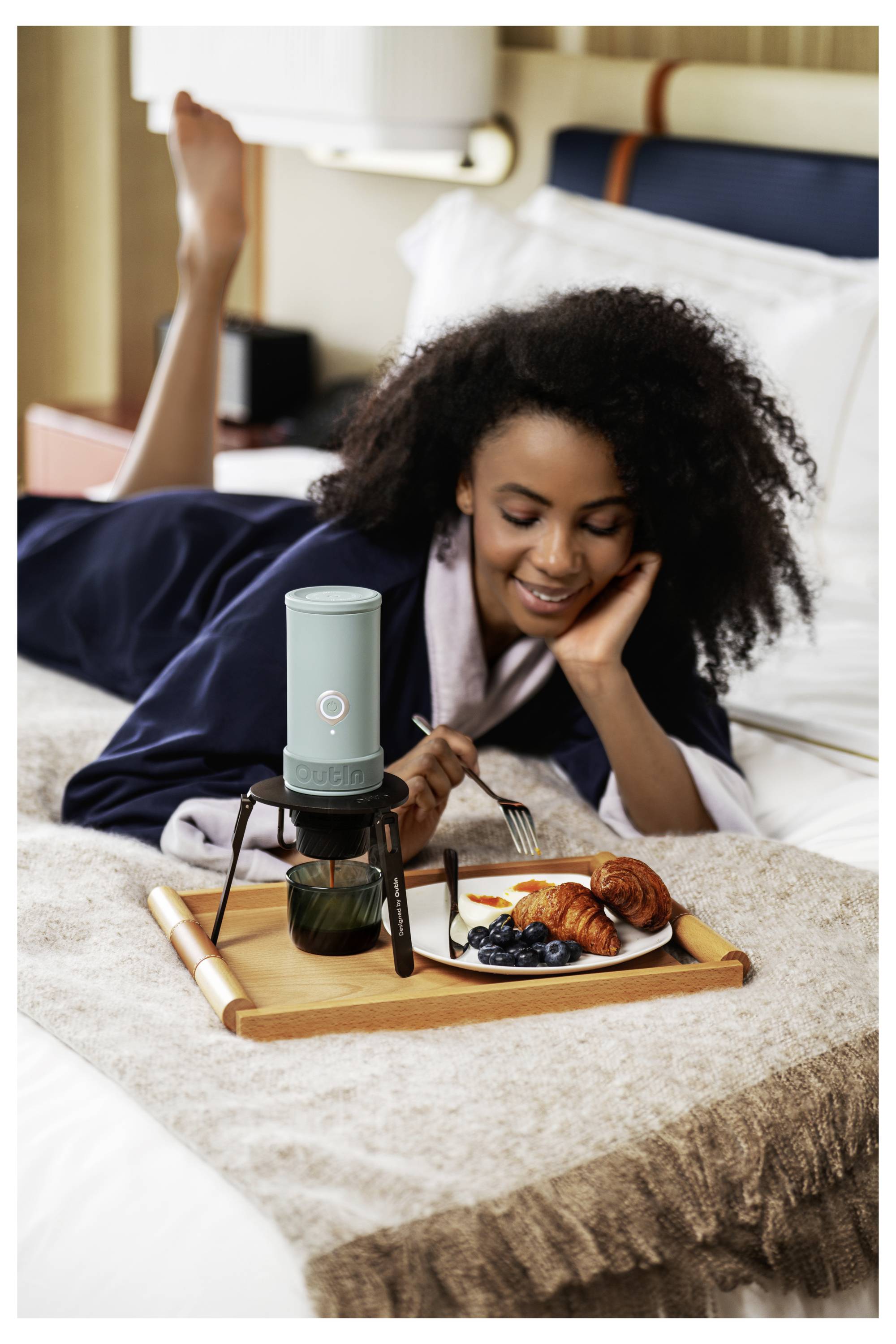 Eine Frau liegt auf einem Bett und genießt Frühstück von einem Tablett mit Kaffee, Croissant, Beeren und einem tragbaren Mikrofon oder Lautsprecher in der Nähe.