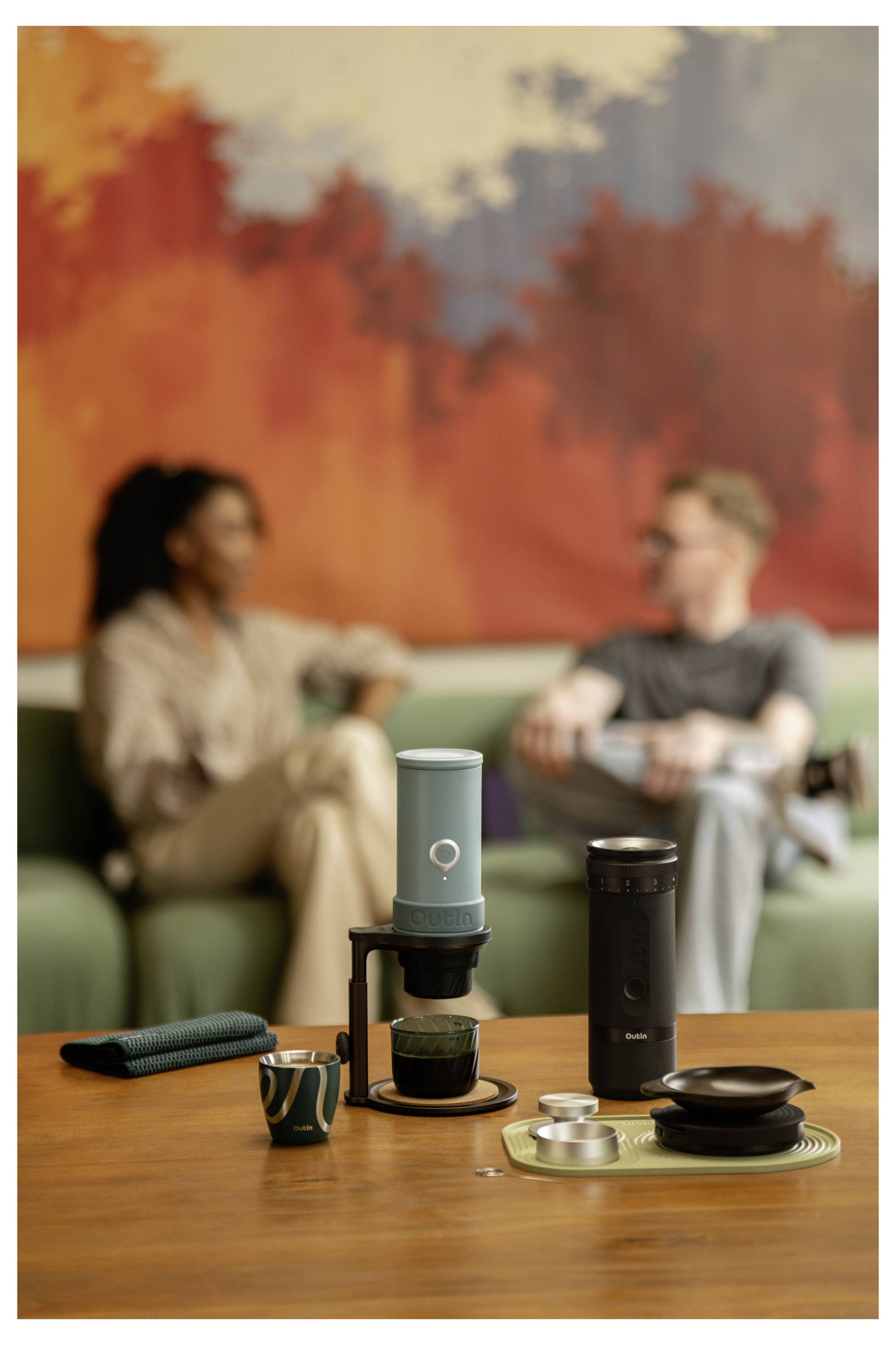 Eine Kaffeezubereitungs-Station auf dem Tisch mit einer Mühle, Tassen und Brühgeräten vor zwei Personen, die auf einem Sofa miteinander plaudern, mit abstrakter Wandkunst hinter ihnen.