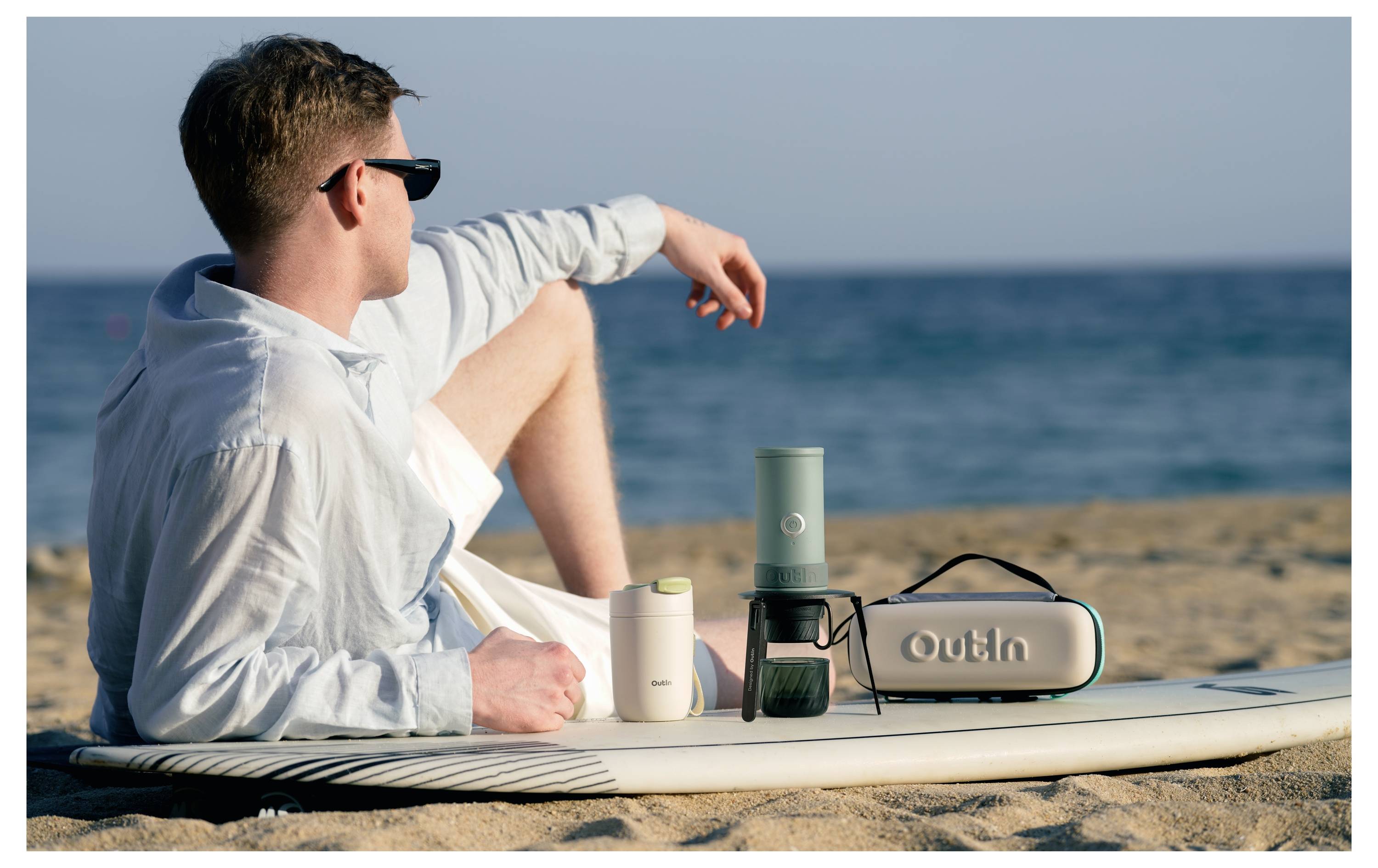 Ein Mann entspannt sich am Strand mit einem Surfbrett und blickt auf den Ozean neben einer tragbaren "Outin"-Kaffeemaschine und Reisebechern.