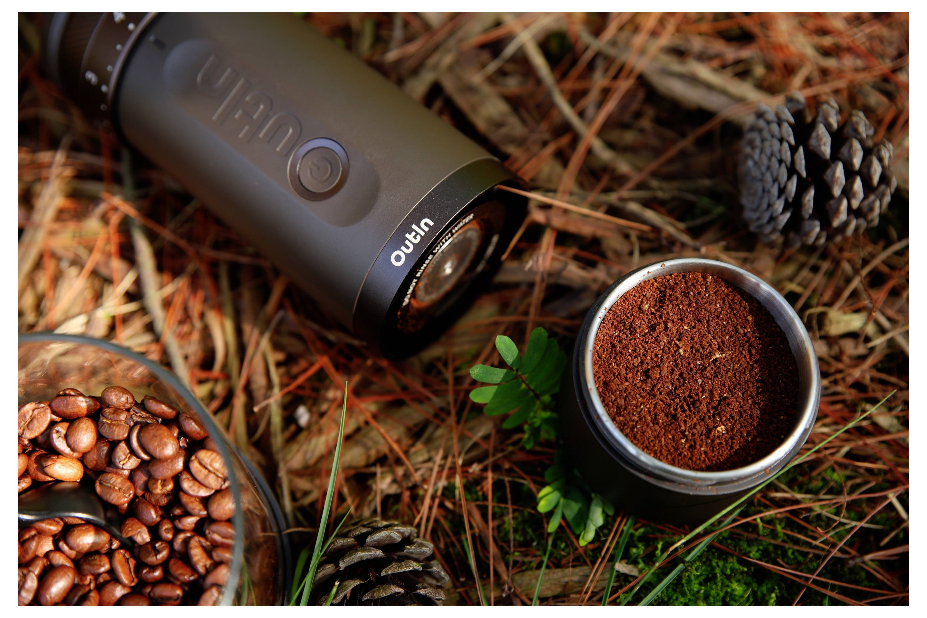 Nahaufnahme einer tragbaren Kaffeemühle mit Kaffeebohnen in einer Schüssel und frisch gemahlenim Kaffee in einer Tasse auf Kiefernnadeln und Gras im Freien.