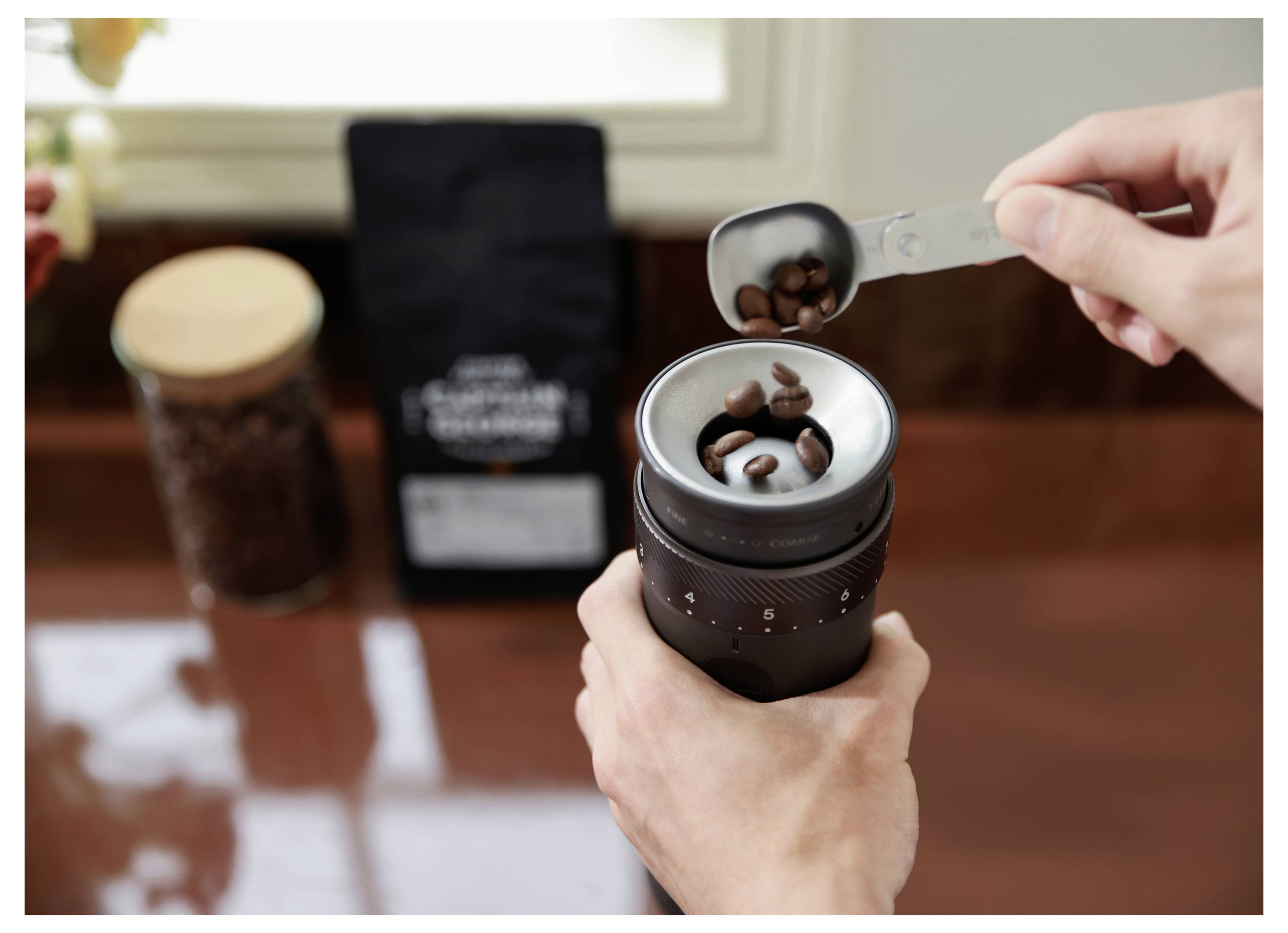Hände gießen Kaffeebohnen in eine manuelle Mühle auf einem Tisch, mit einer Tüte und einem Glas mit Bohnen im Hintergrund.