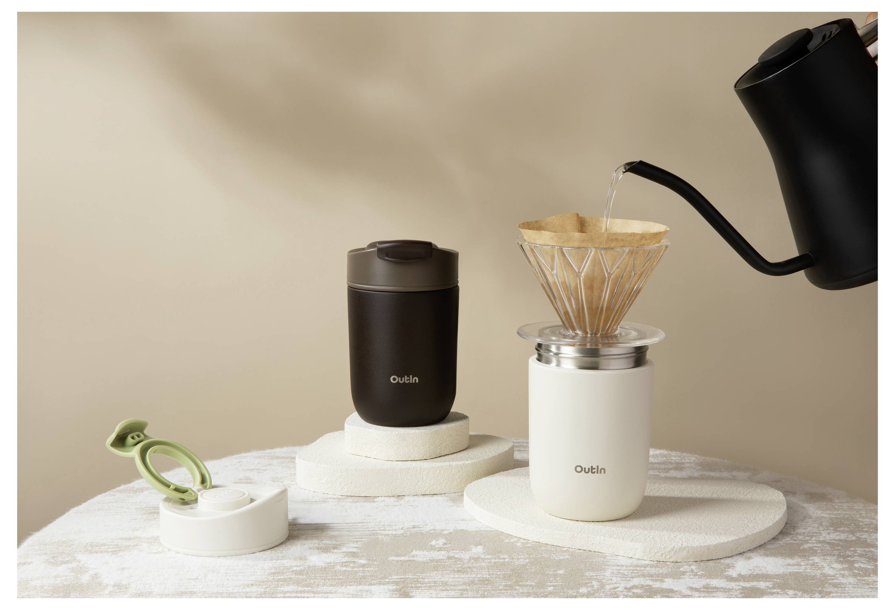 Ein schwarzer Wasserkocher gießt Wasser in einen Kaffeefilter auf einer weißen Tasse neben einer dunklen tragbaren Mühle und einer grünen Klammer auf einem strukturierten Tisch.