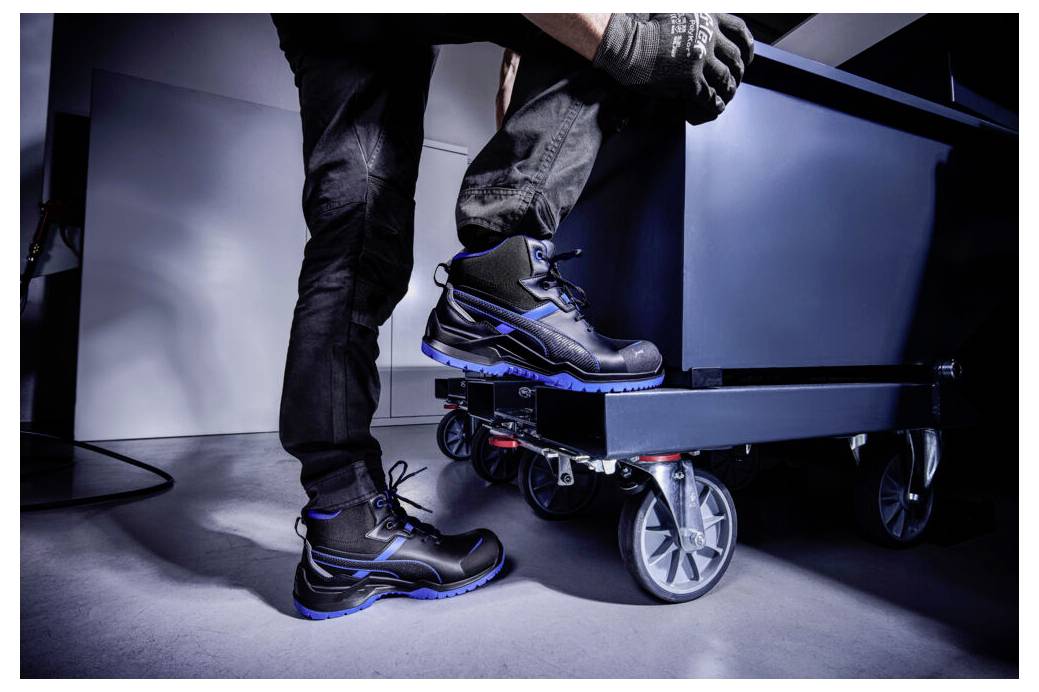 Eine Person, die schwarze Arbeitsstiefel trägt, tritt auf einen kleinen fahrbaren Wagen oder Transportwagen in einer studioähnlichen Umgebung.