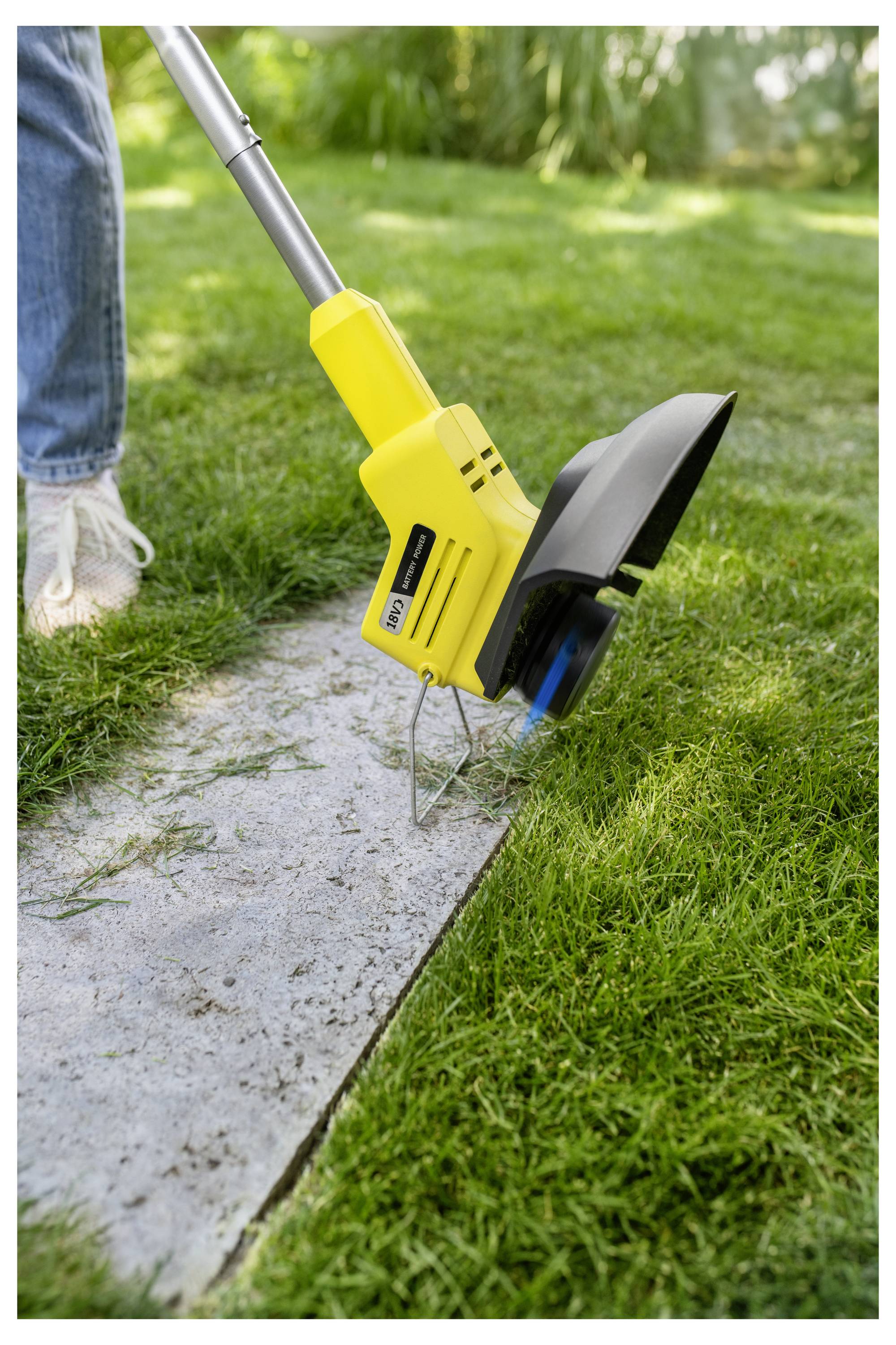 Nahaufnahme eines gelben elektrischen Rasenkantenschneiders, der Gras entlang eines Betonwegs schneidet, mit einem sichtbaren Bein einer Person auf der linken Seite.