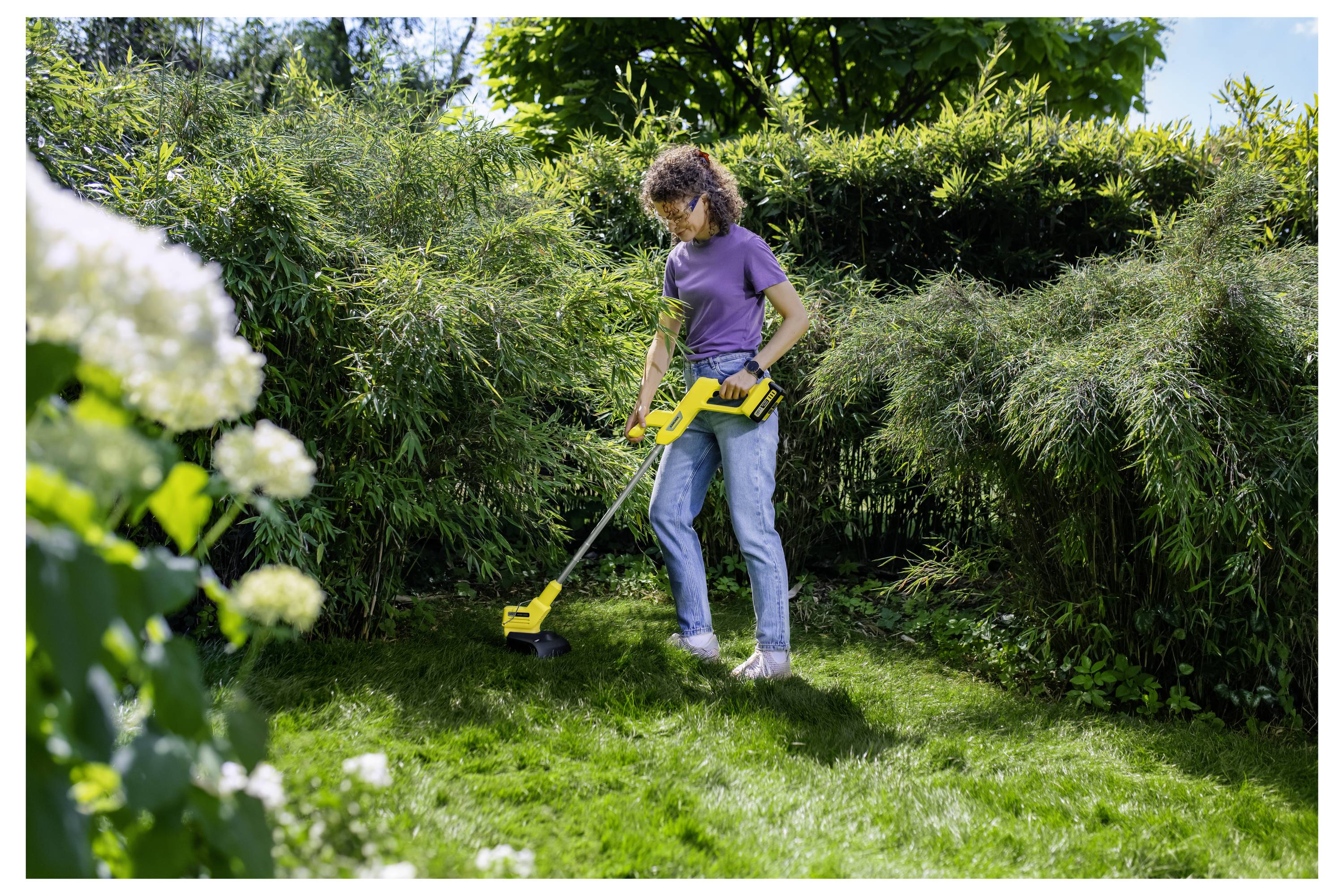 Eine Frau, die mit einem Fadentrimmer Gras in einem üppigen Hinterhofgarten schneidet, mit hohen Hecken und Blumen im Vordergrund.