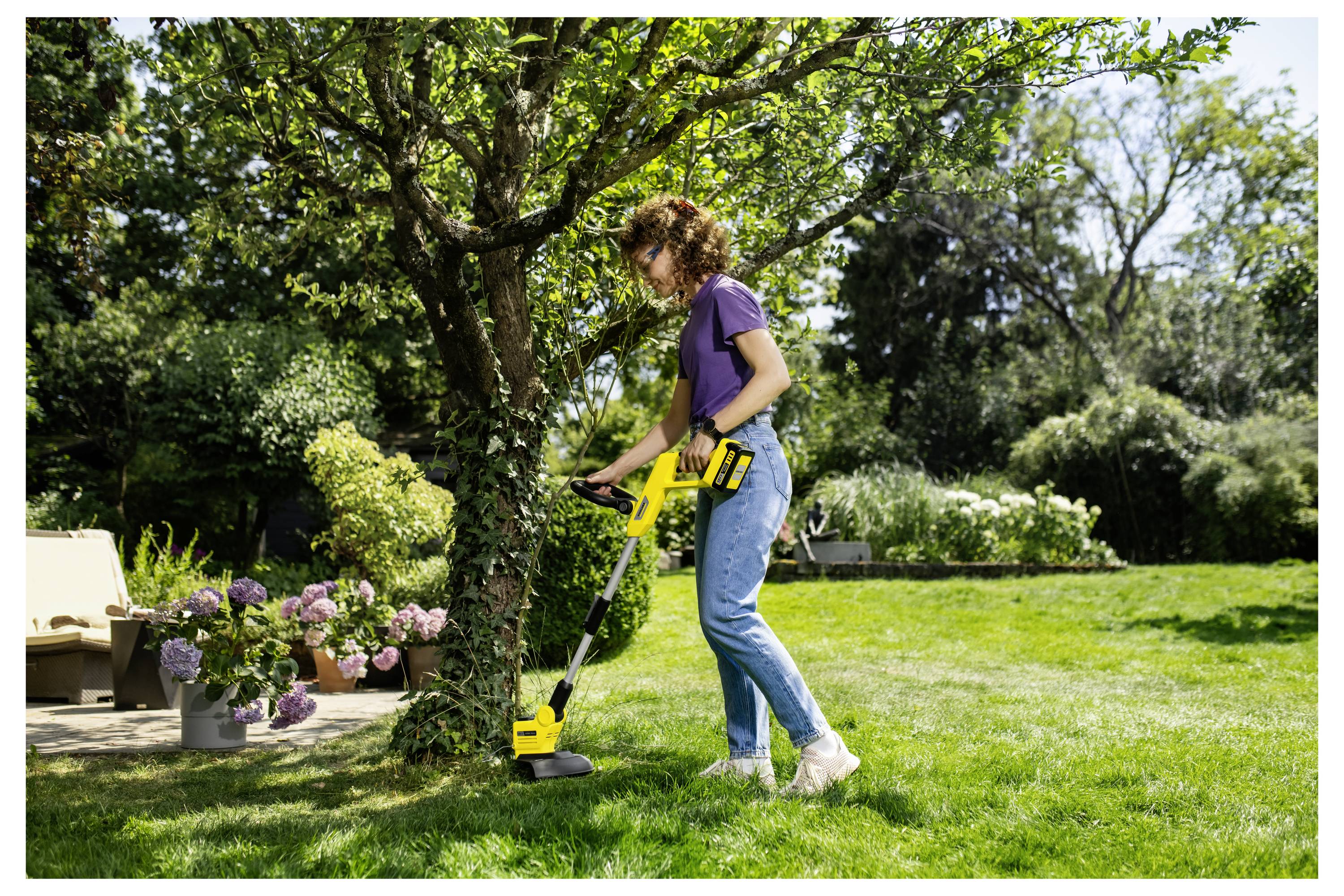Frau, die einen gelben kabellosen Rasenkantentrimer verwendet, um Gras um einen Baum in einem sonnigen Garten im Hinterhof zu kanten.