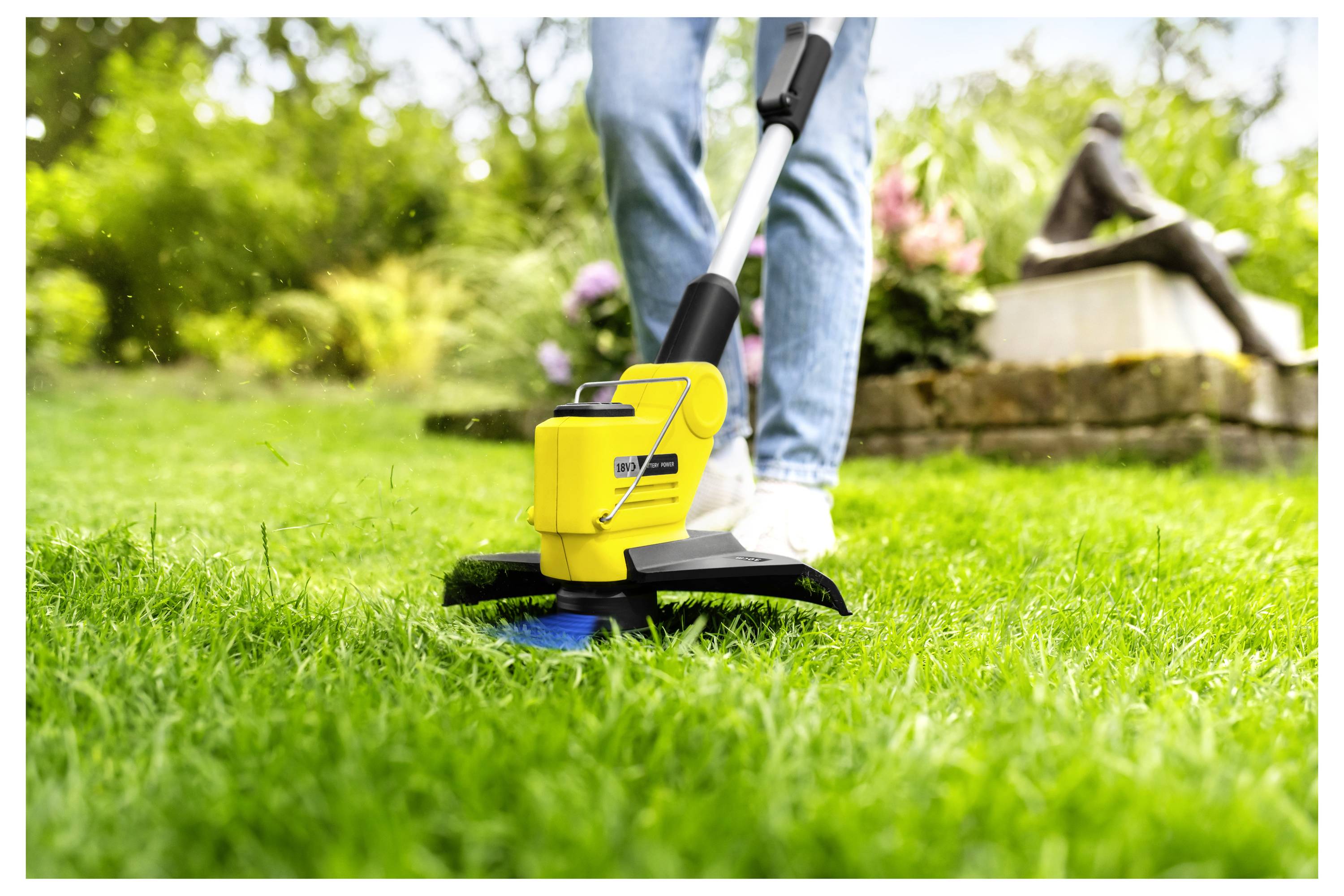 Eine Person benutzt einen gelben elektrischen Rasentrimmer, um Gras in einem Garten zu schneiden, wobei nur ihre Beine sichtbar sind.