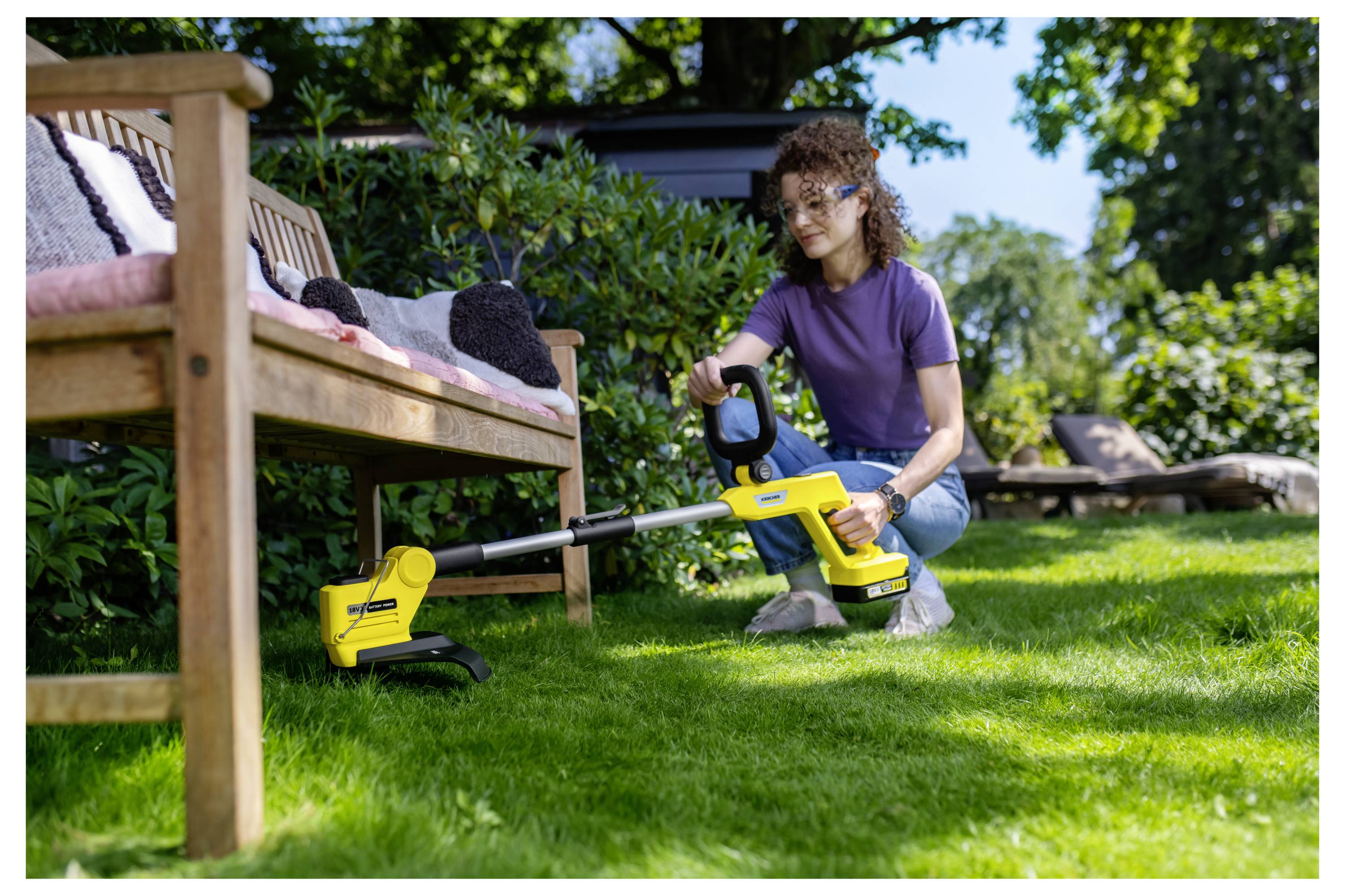 Frau kniet auf einem Rasen und benutzt einen gelben kabellosen Rasentrimmmer neben einer Holzbank in einem sonnigen Garten.