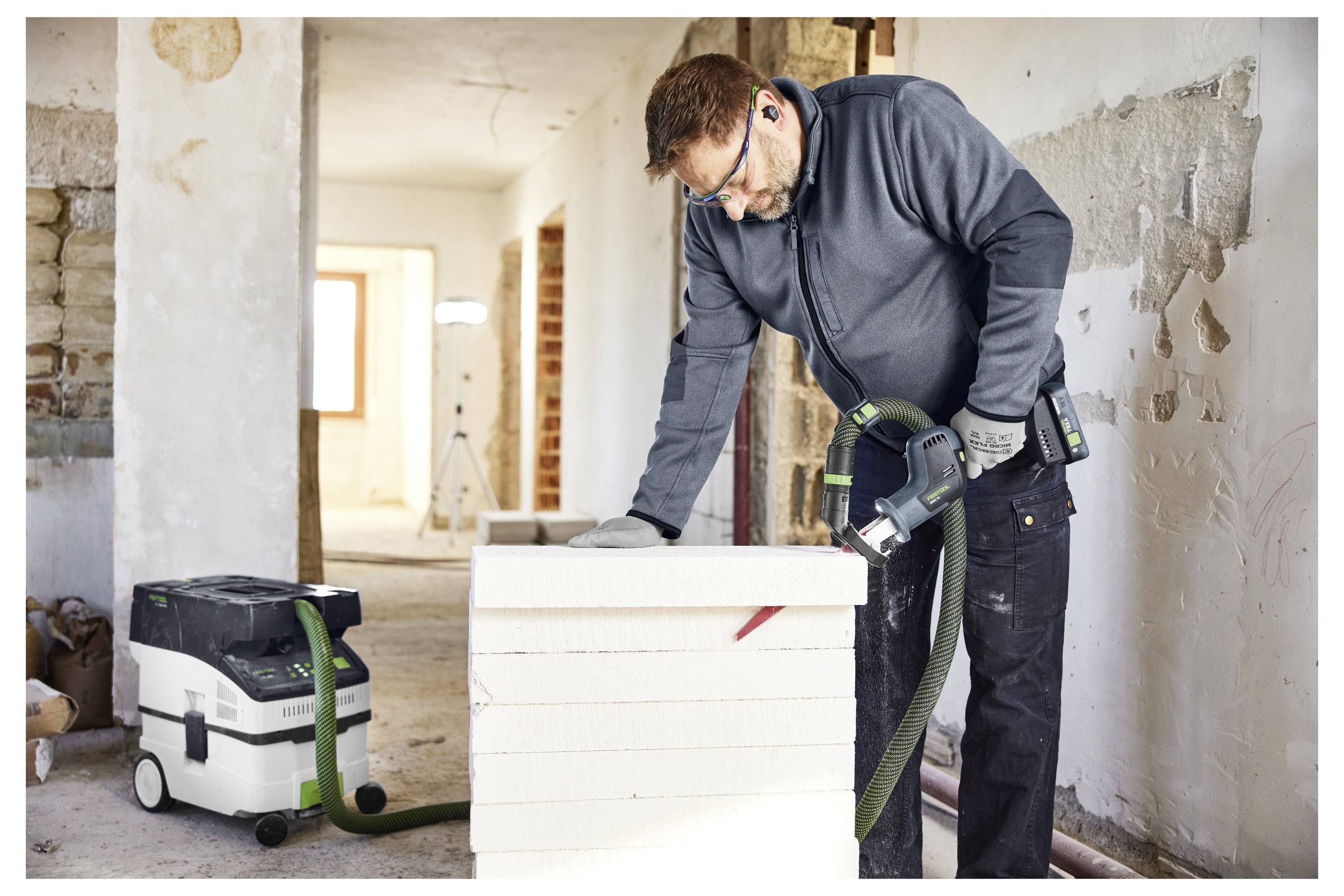 Ein Arbeiter in einem teilweise renovierten Raum verwendet einen schlauchangeschlossenen Staubsauger auf einem weißen Panel oder Block neben einer Reinigungsmaschine.
