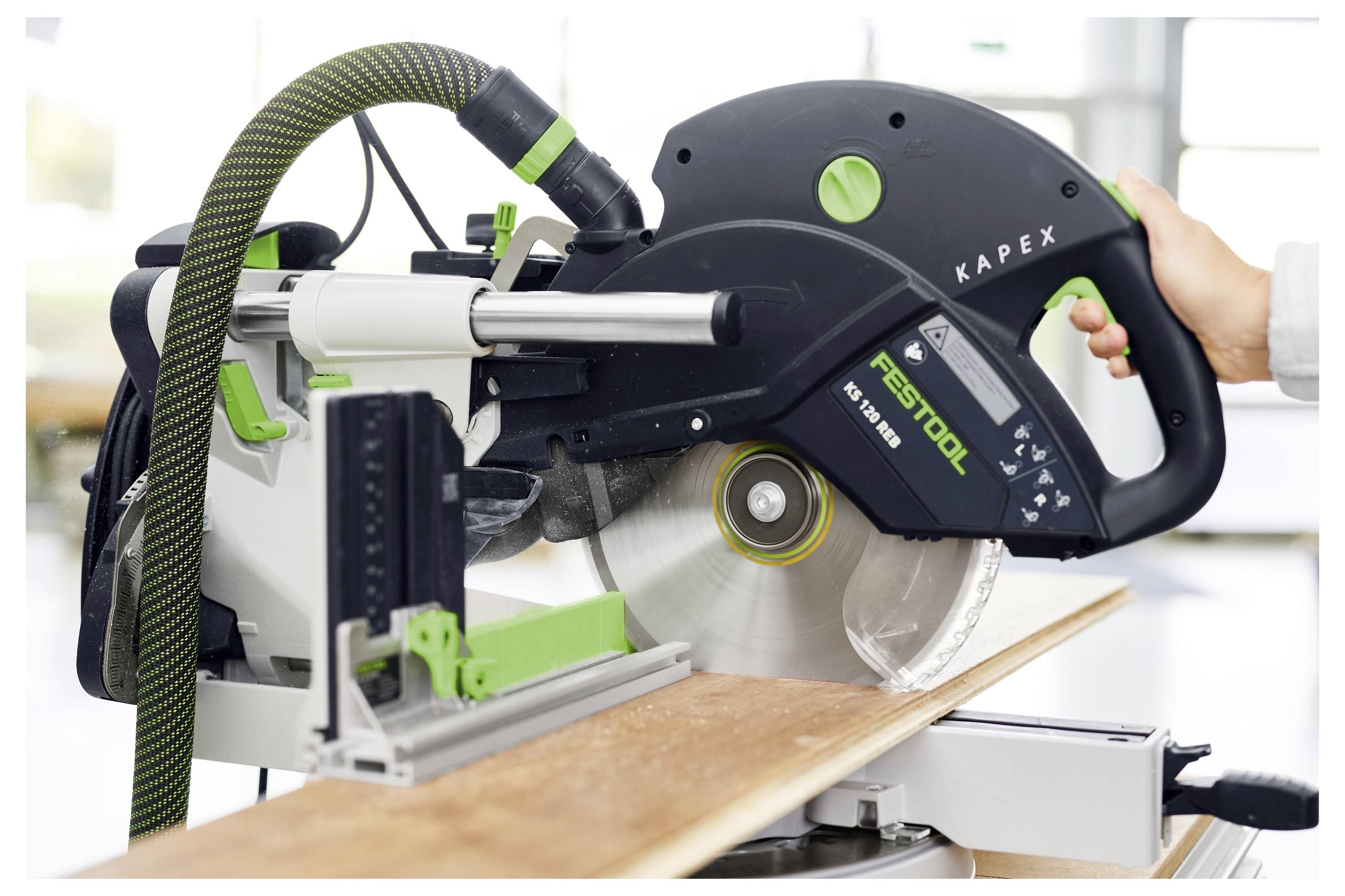 Eine Festool Kapex Gleitgehrungssäge schneidet ein Holzbrett, wobei eine Hand den Griff hält und das Sägeblatt in einer Werkstatt dreht.