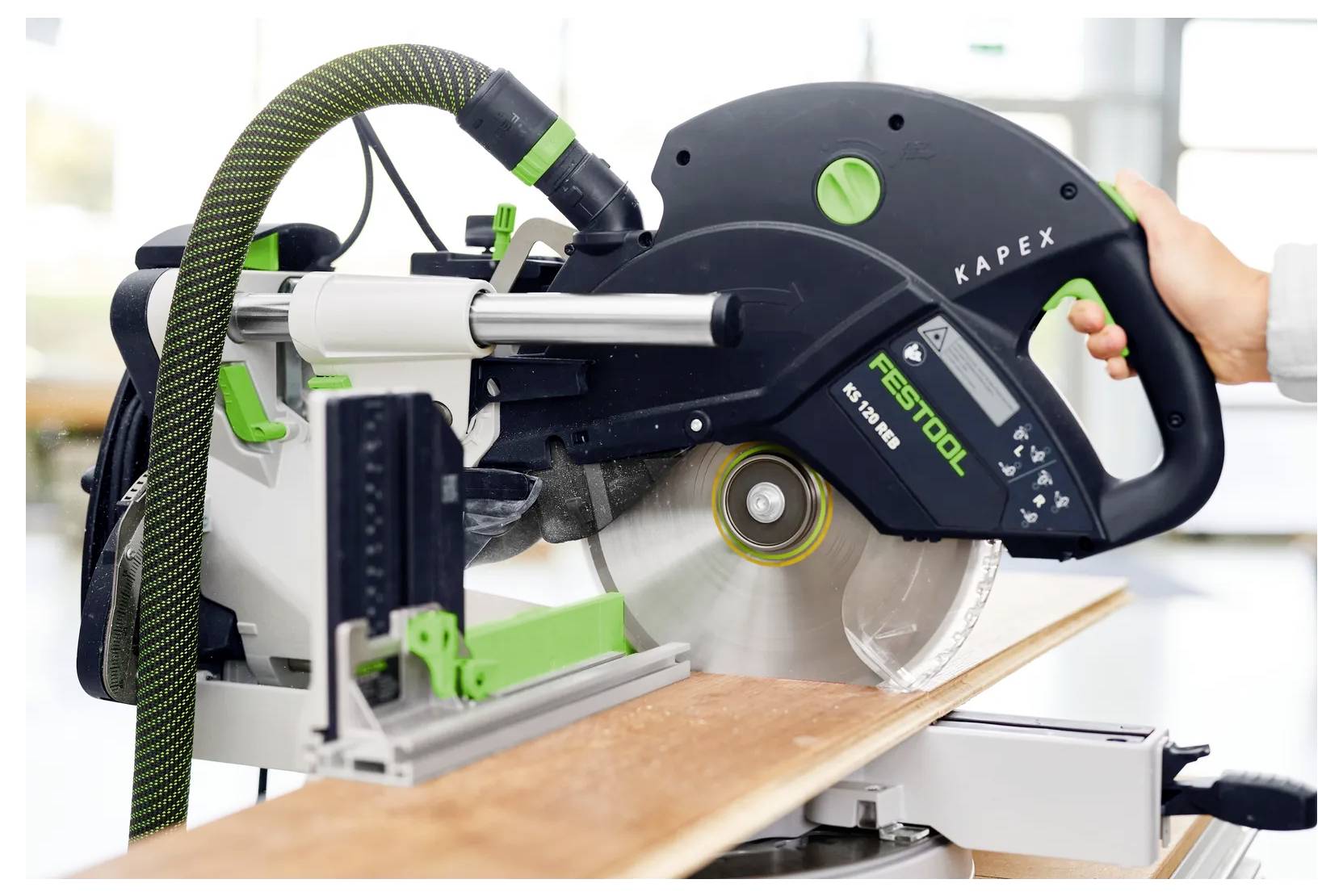 Ein Festool Kapex-Kappschleifer schneidet ein Holzbrett, mit einer Hand, die den Griff hält und die Klinge während des Gebrauchs freigelegt ist.