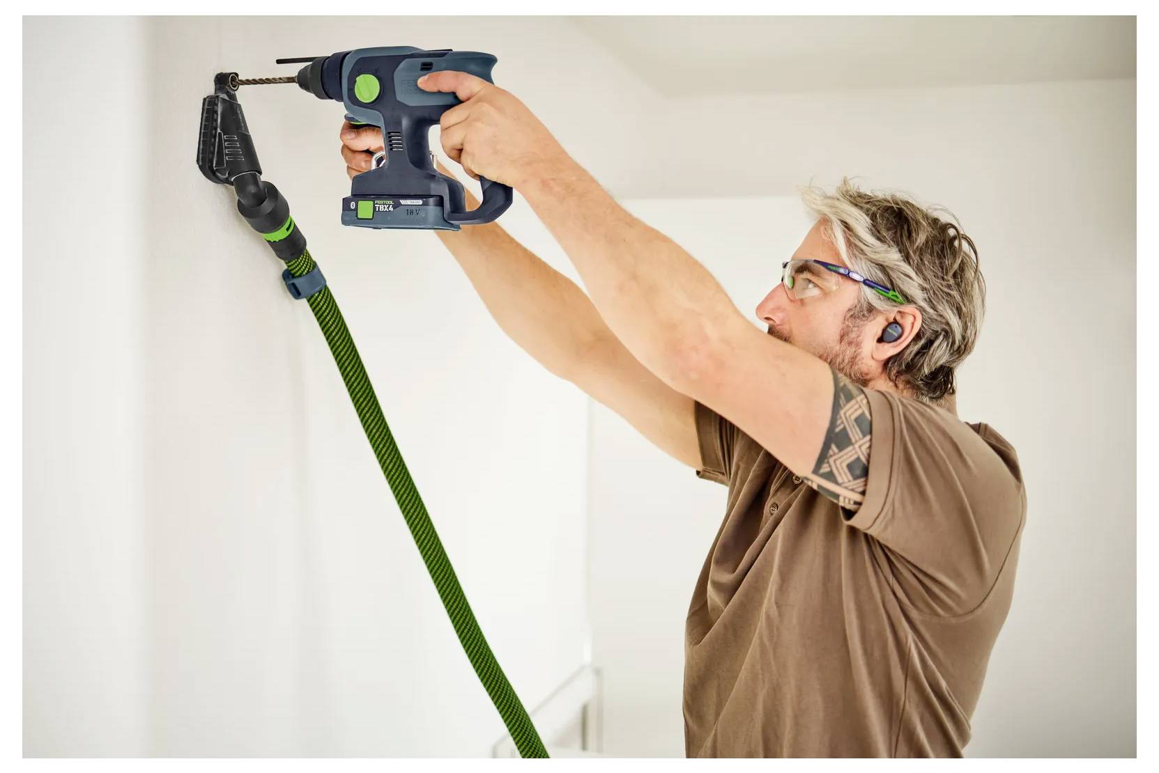 Ein Mann bohrt mit einer schnurgebundenen Bohrmaschine mit angeschlossener Absaugschlauch-Vorrichtung in eine Wand in der Nähe der Decke, wobei er eine Schutzbrille und Gehörschutzstöpsel trägt.