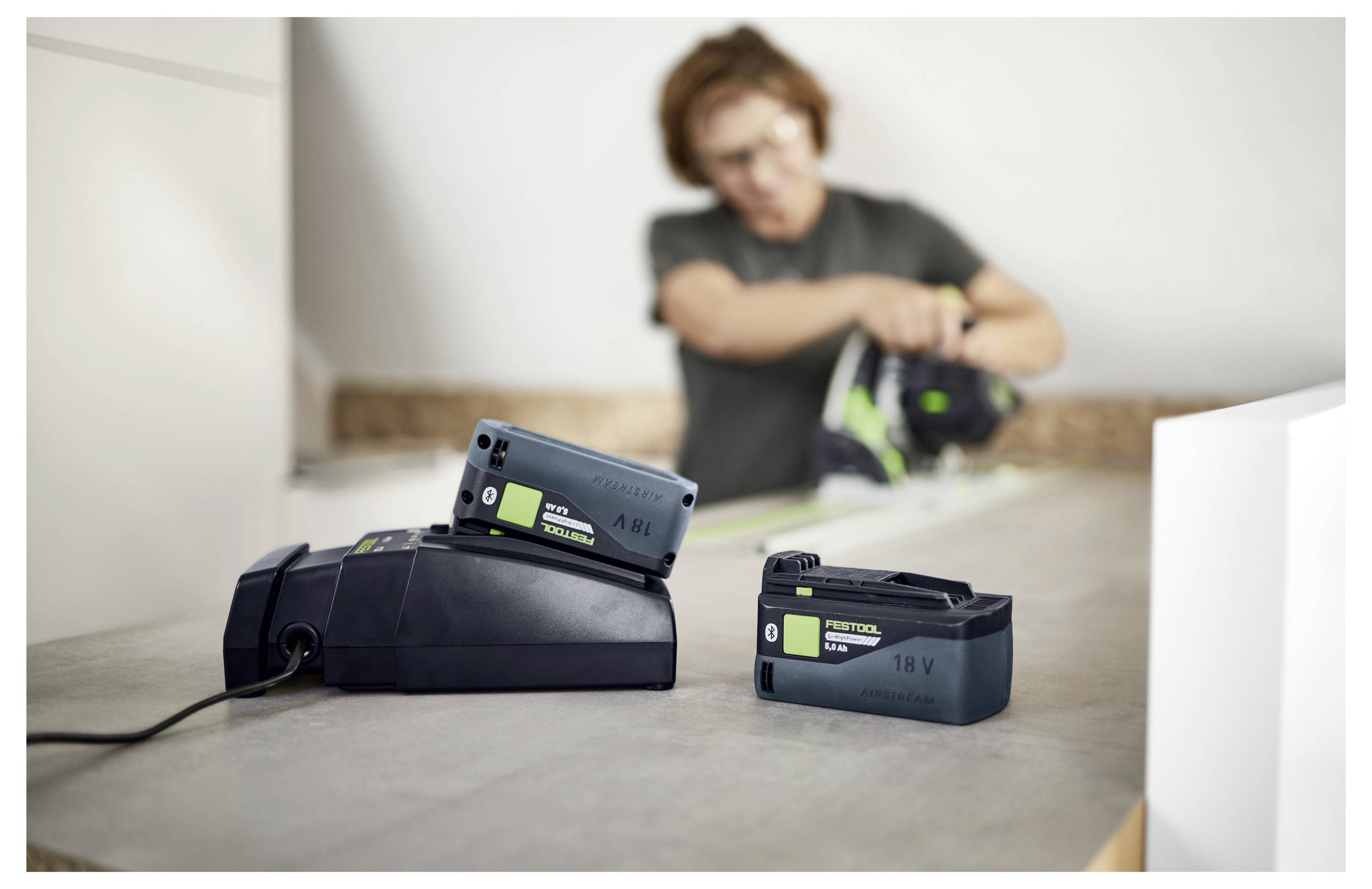 Zwei kabellose Werkzeugbatterien auf einem Ladegerät im Vordergrund, mit einer unscharfen Person, die im Hintergrund arbeitet.