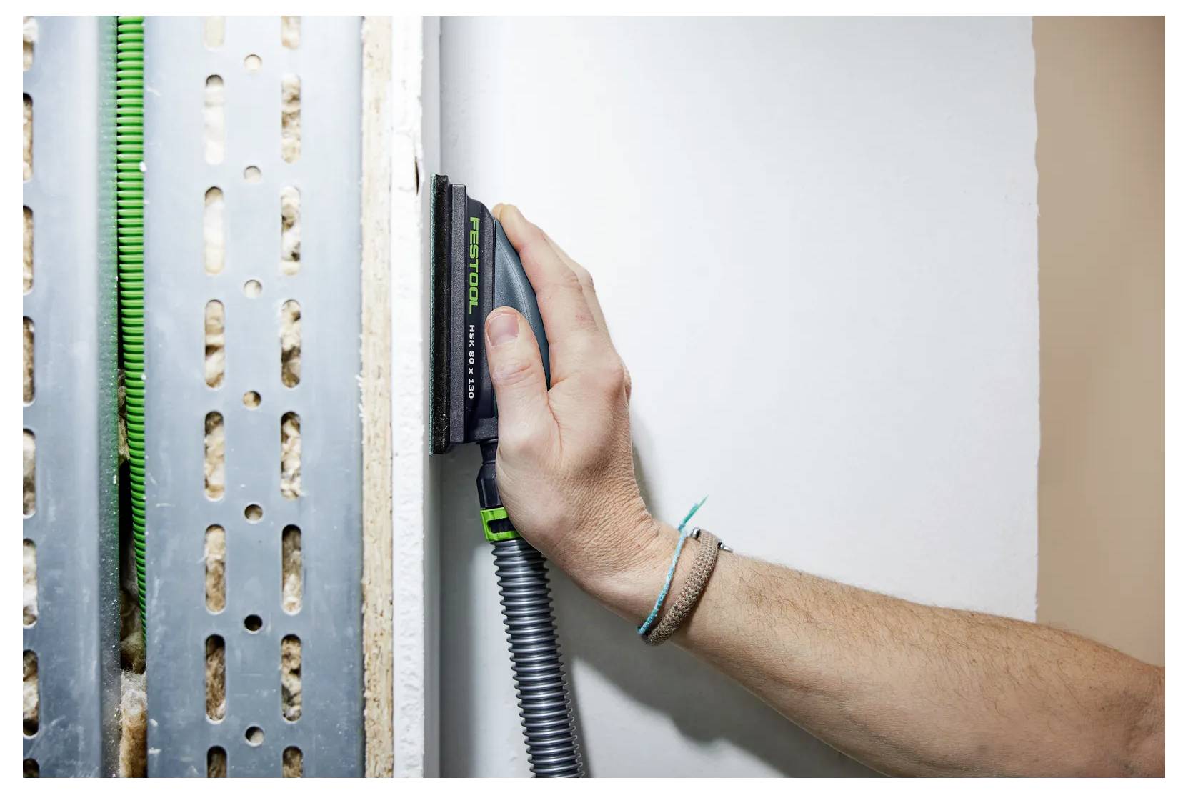 Nahaufnahme einer Hand, die einen Bolzensucher an einer Wand neben freiliegendem Metallrahmen verwendet, um einen versteckten Bolzen oder ein Rohr zu lokalisieren.