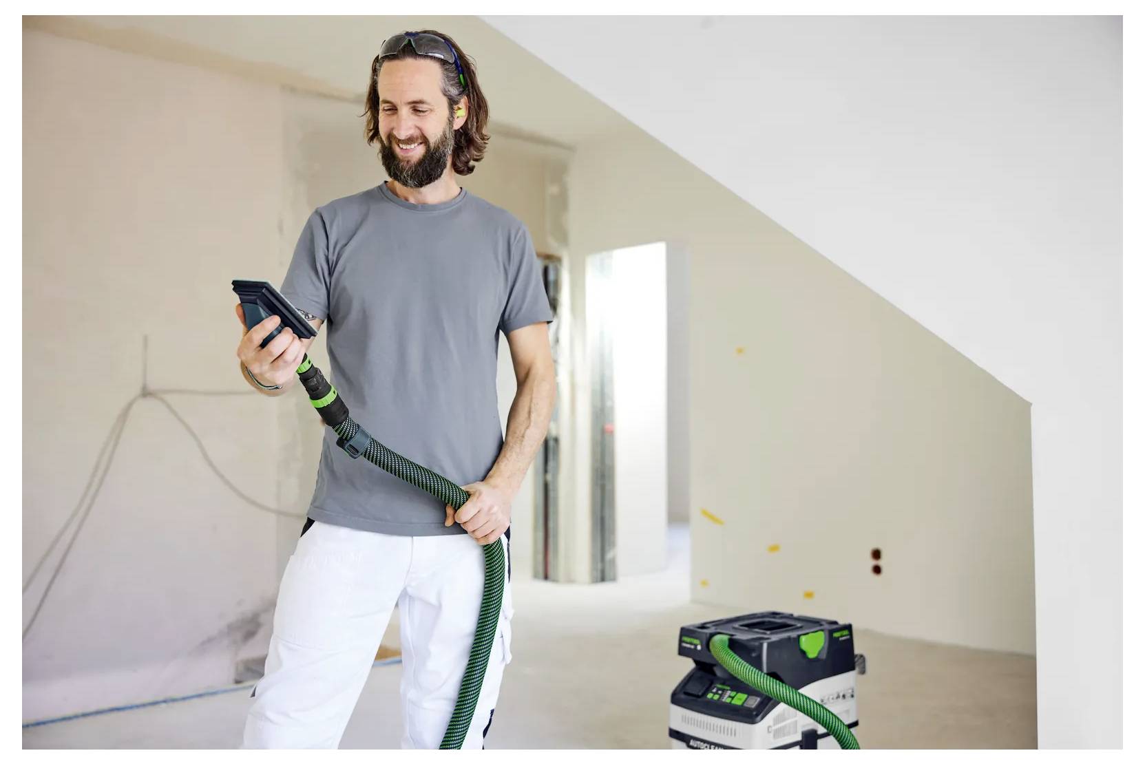 Lächelnder Mann in einem grauen Hemd und weißer Hose hält einen Elektrowerkzeugschlauch in einem unfertigen Raum, mit einem tragbaren Staubsauger oder einer Absauganlage in der Nähe.