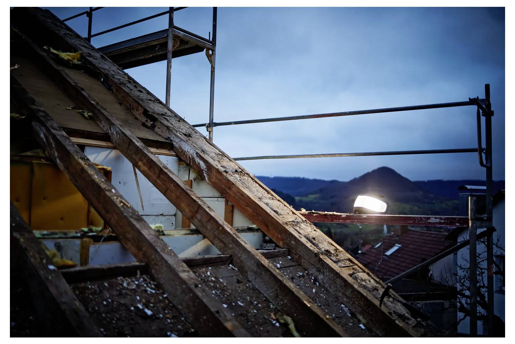 Verfallende Holzdachbalken unter Gerüsten in der Dämmerung, mit einem hellen Arbeitslicht und entfernten Bergen im Hintergrund.