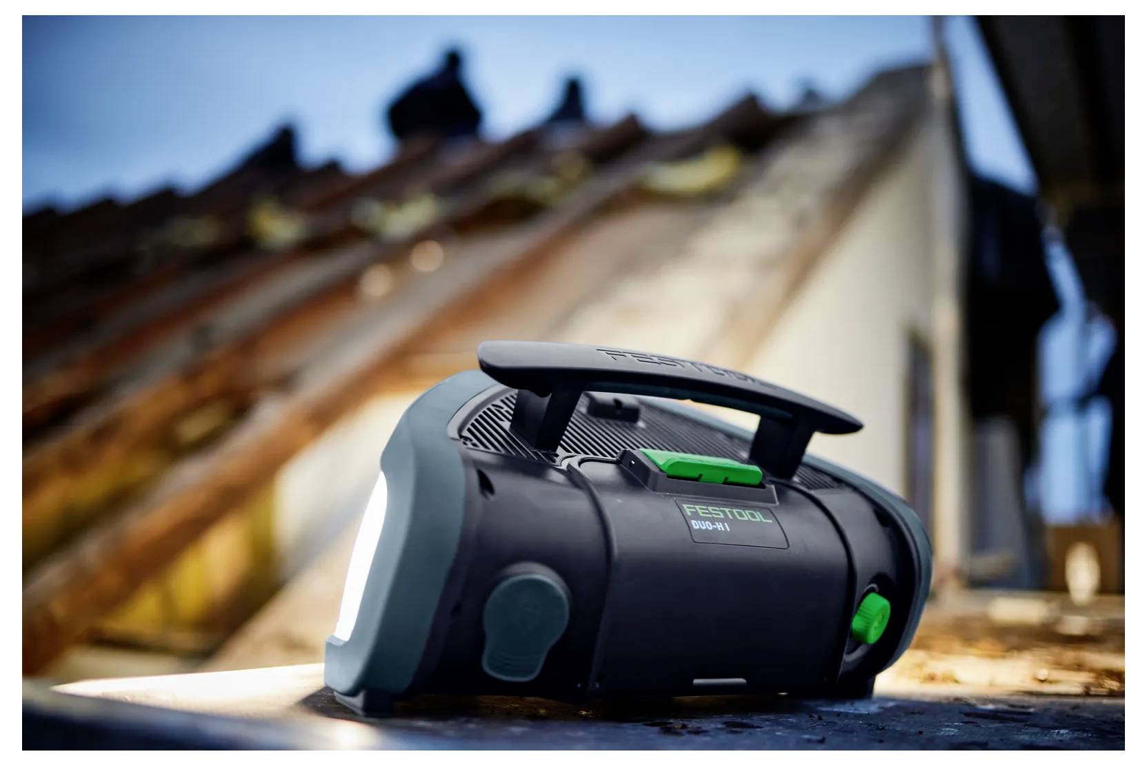 Tragbares schwarzes Festool Akku-Funkgerät/Werkzeugakku auf einer Arbeitsfläche, mit unscharfem Dach und Arbeiter im Hintergrund.