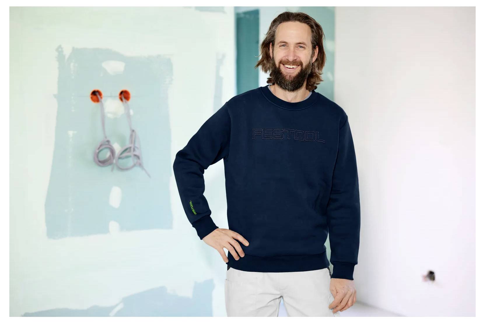 Lächelnder Mann in einem dunklen Pullover steht in einem Raum mit einer hellblau gestrichenen Wand, auf der freiliegende elektrische Leitungen und unvollendete Innenarbeiten sichtbar sind.
