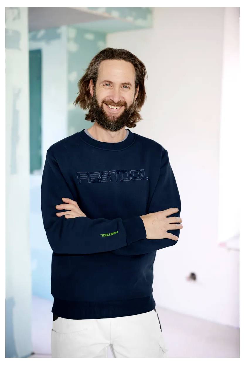 Lächelnder bärtiger Mann in einem dunklen Sweatshirt und weißer Hose steht mit verschränkten Armen im Innenbereich.