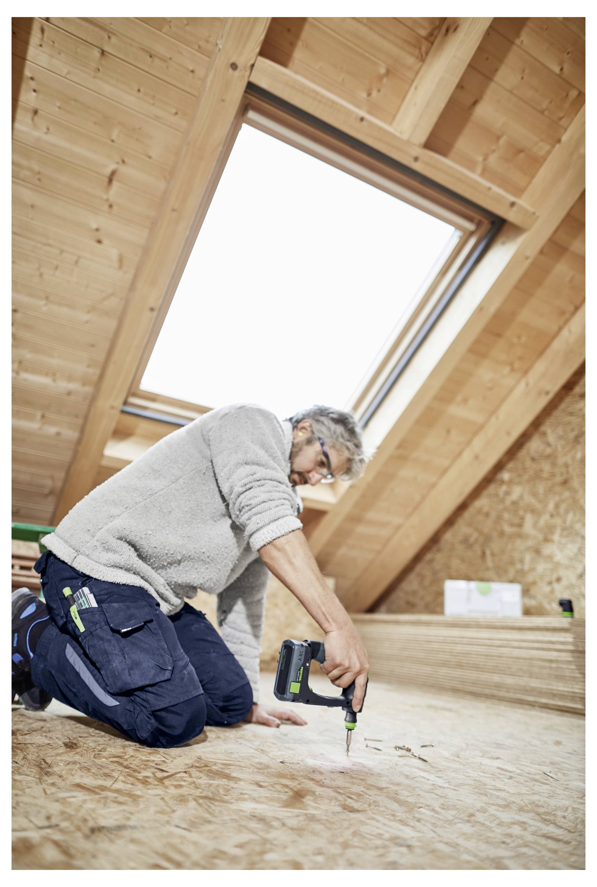 Ein Zimmermann kniet auf einem Holzboden auf dem Dachboden und bohrt in das Sperrholz unter einem Dachfenster.