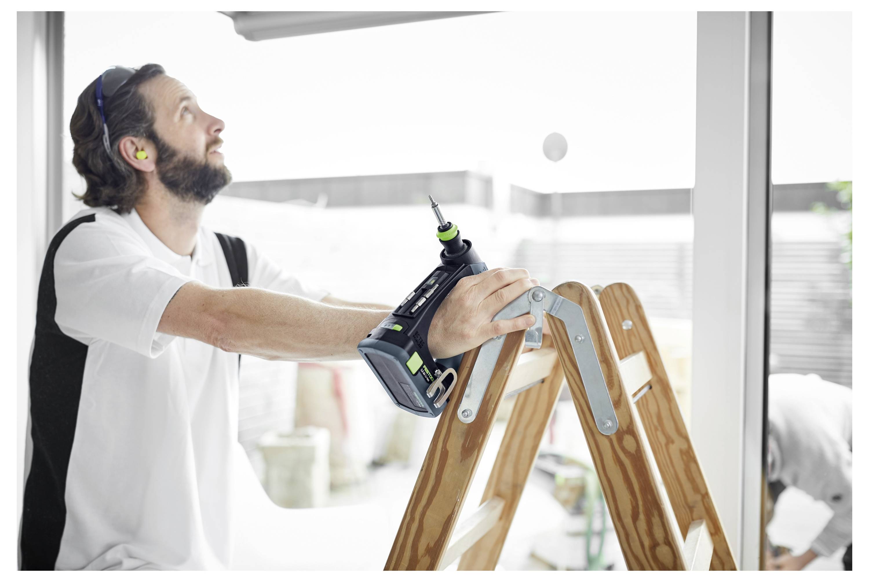 Ein Mann mit Gehörschutz bedient einen Bohrer, während er auf einer Stehleiter steht und Heimwerkerreparaturen im Innenbereich durchführt.
