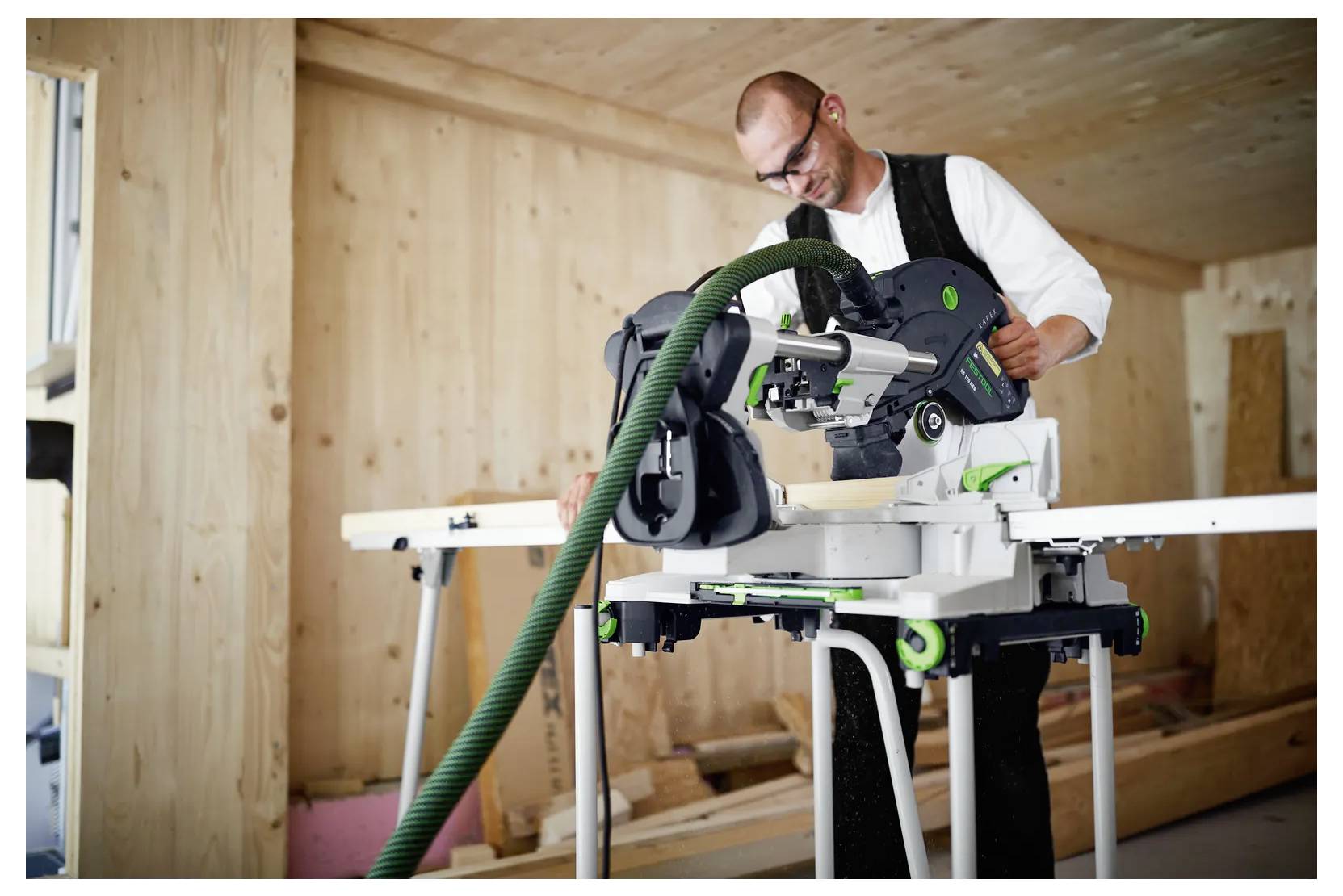 Ein Mann benutzt eine Gehrungssäge, um Holz in einem unfertigen Holzzimmer zu schneiden, mit angeschlossener Staubabsaugschlauch.