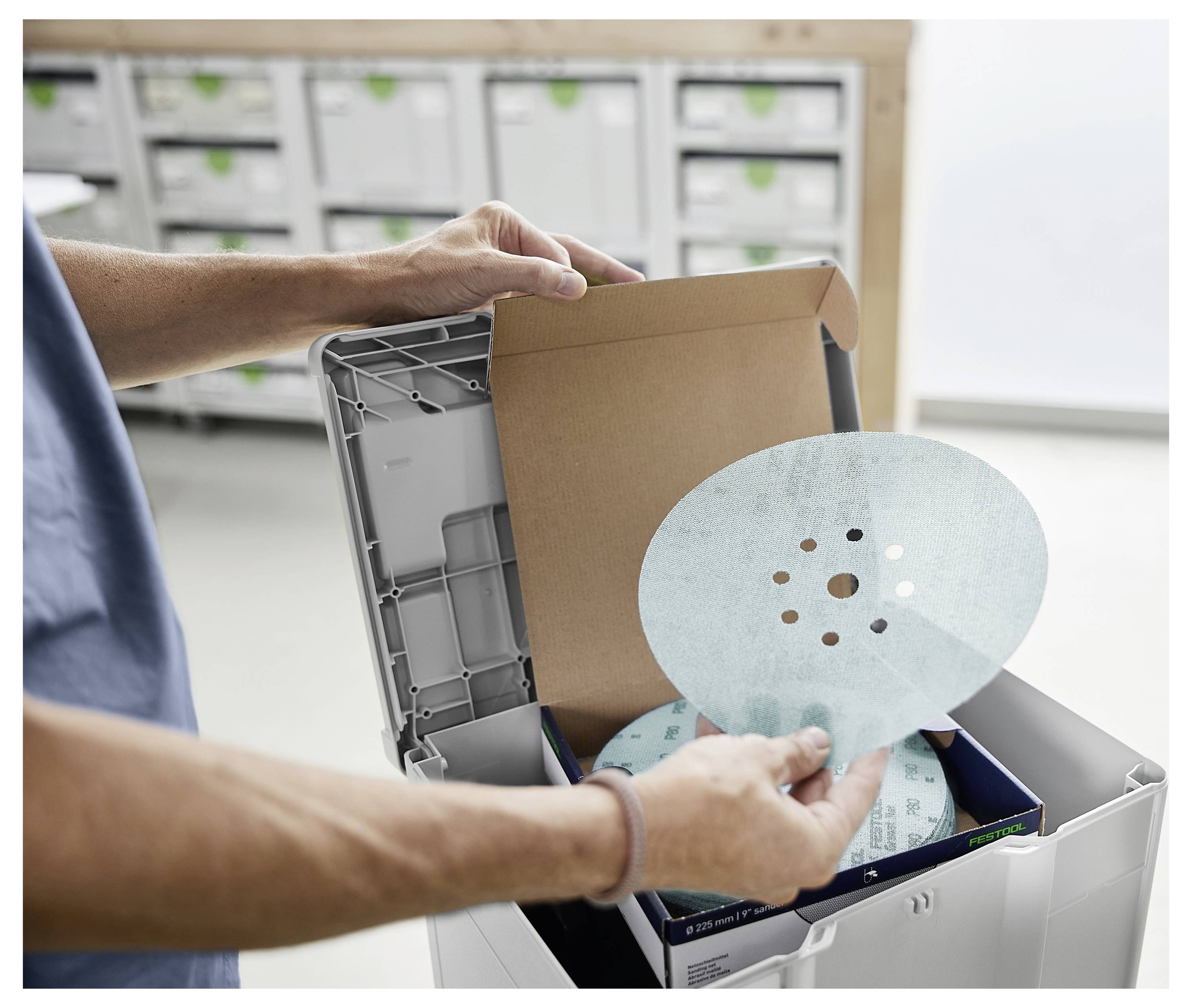 Eine Person packt ein Paket in einer Werkstatt aus und entfernt eine kreisförmige Schleifscheibe aus einer Kartoneinlage.