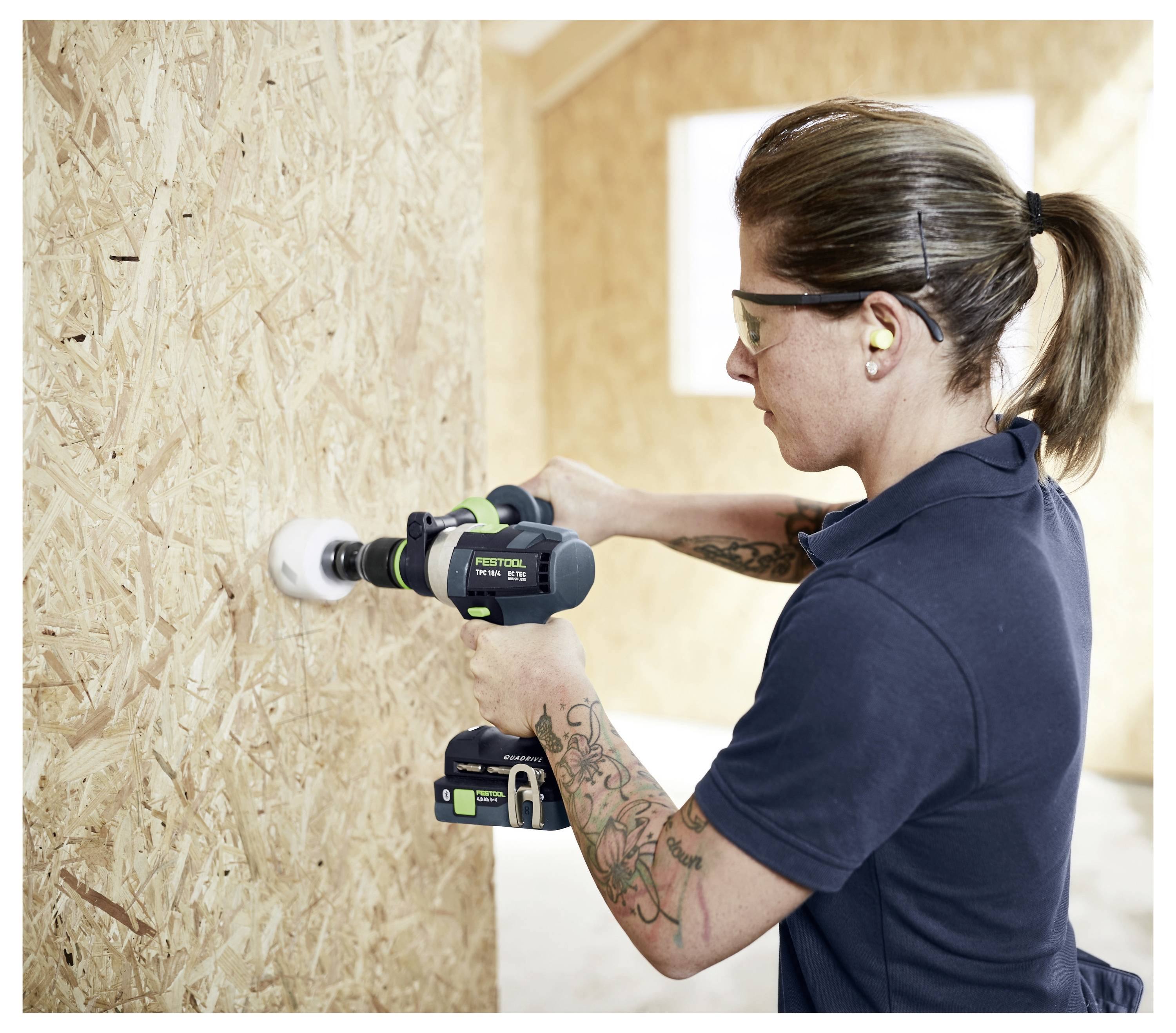 Frau mit Schutzbrille bohrt mit einer Elektrobohrmaschine ein Loch in eine Sperrholzwand auf einer Baustelle.