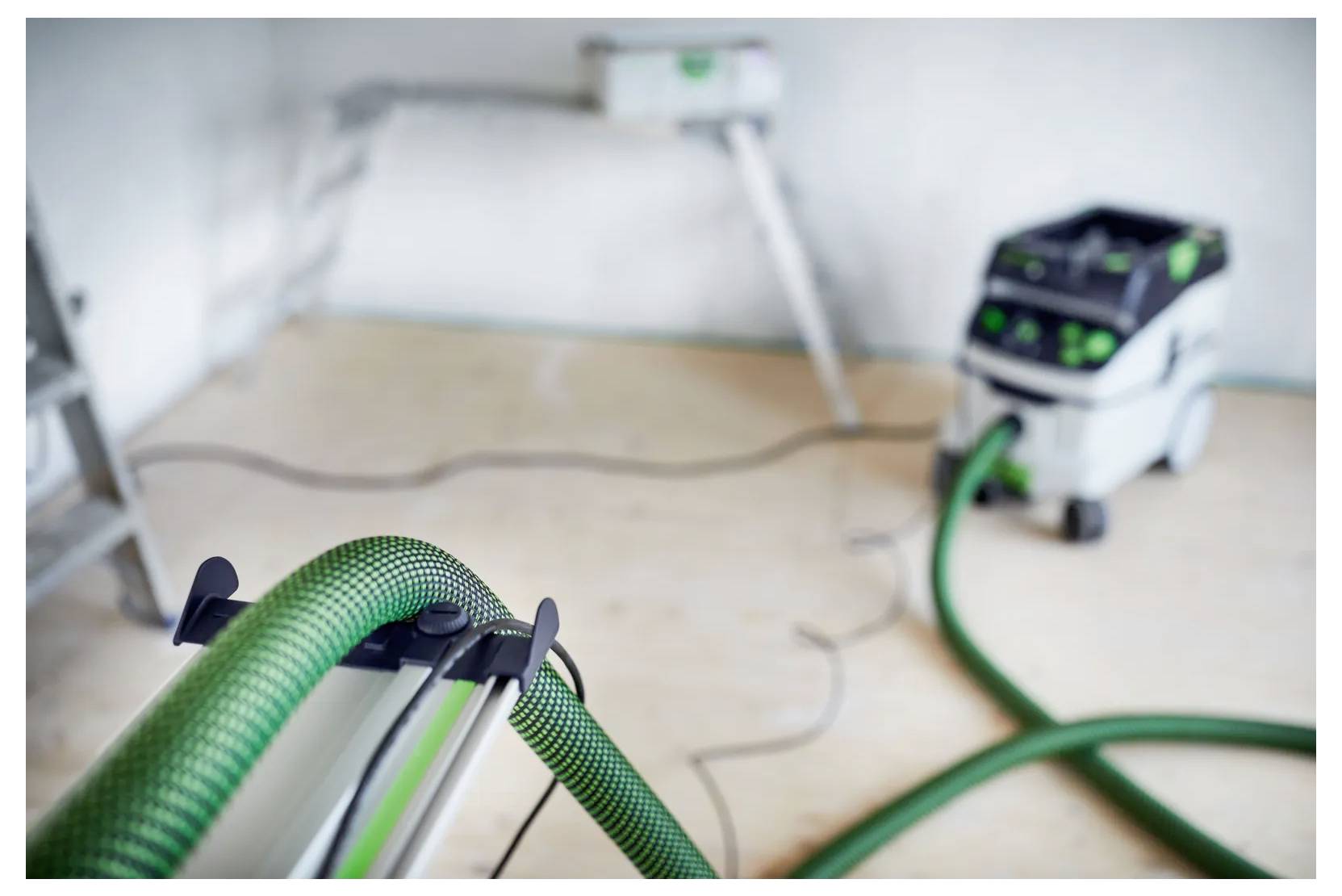 Elektroschliff mit grünem Schlauch in einem Renovierungszimmer, mit Schleifausrüstung im Vordergrund und einem Staubabscheider im Hintergrund.