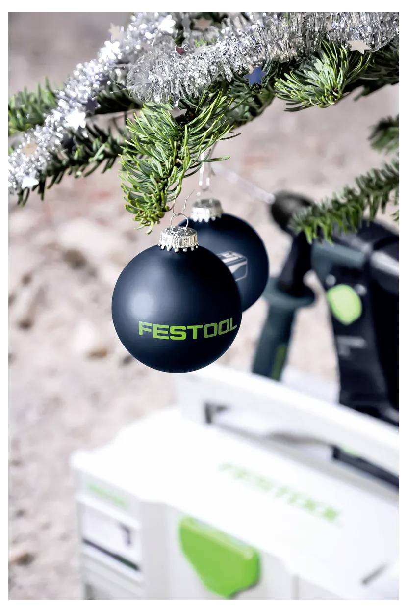 Schwarzes Festool-Ornament hängend an einem Weihnachtsbaumzweig, mit einem unscharfen Festool-Werkzeugkoffer im Hintergrund.